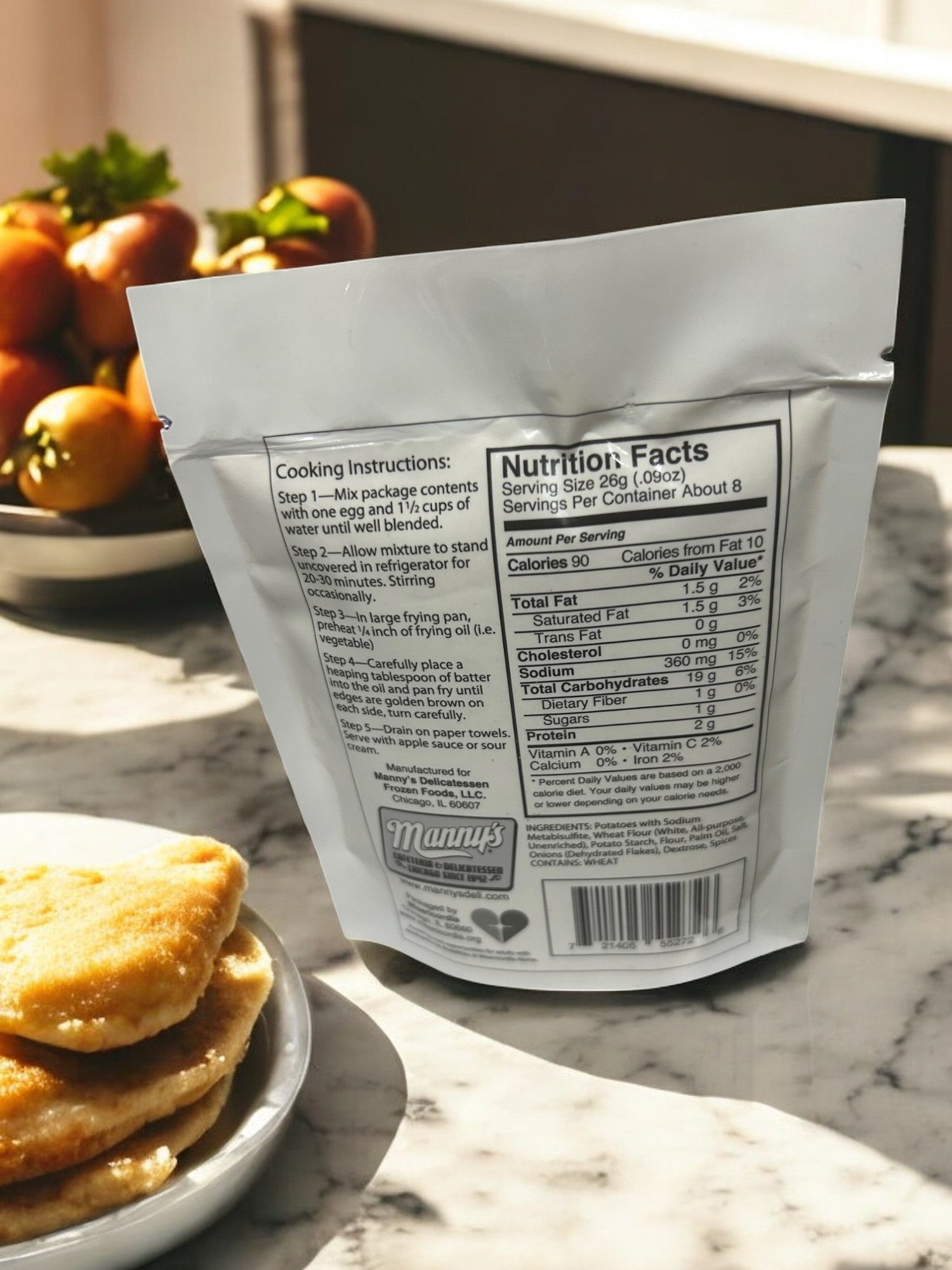 A package of Manny's Deli Potato Pancake Mix-1 Count sits on a kitchen counter, showing cooking instructions and nutritional info. Nearby, tomatoes and bread slices rest on plates as sunlight casts shadows on the marble surface.