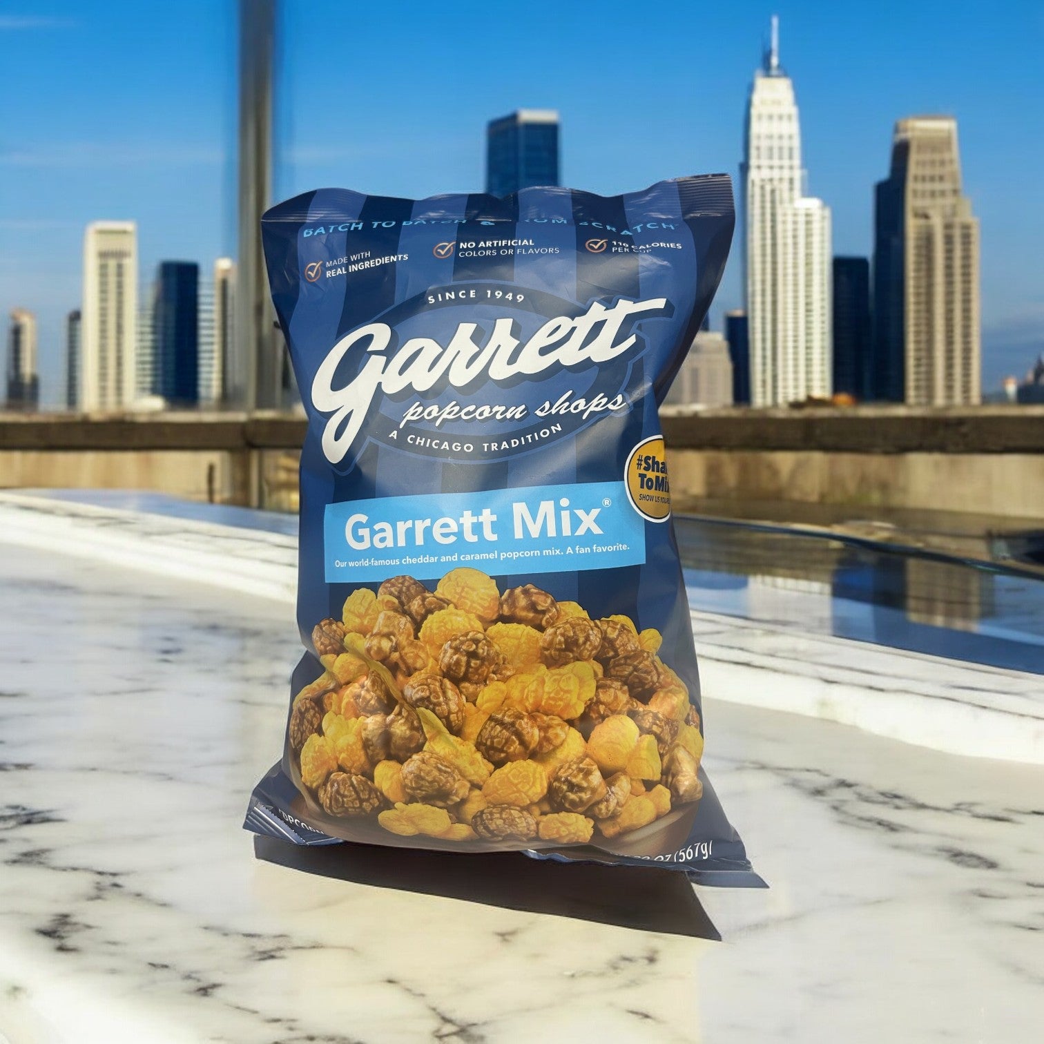 A 20 oz. bag of Garrett Popcorn's Cheddar and Caramel Mix sits on a marble surface, the handcrafted popcorn shining against a vibrant city skyline, capturing the essence of Garrett Popcorn Shops.