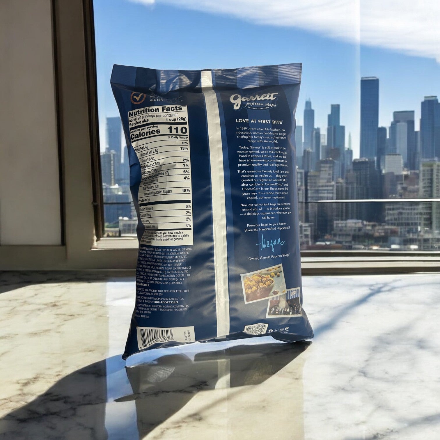 A bag of Garrett Popcorn Cheddar and Caramel Garrett Mix Popcorn-3 Count by Garrett sits on a marble counter, with the city skyline visible through the window in the background.