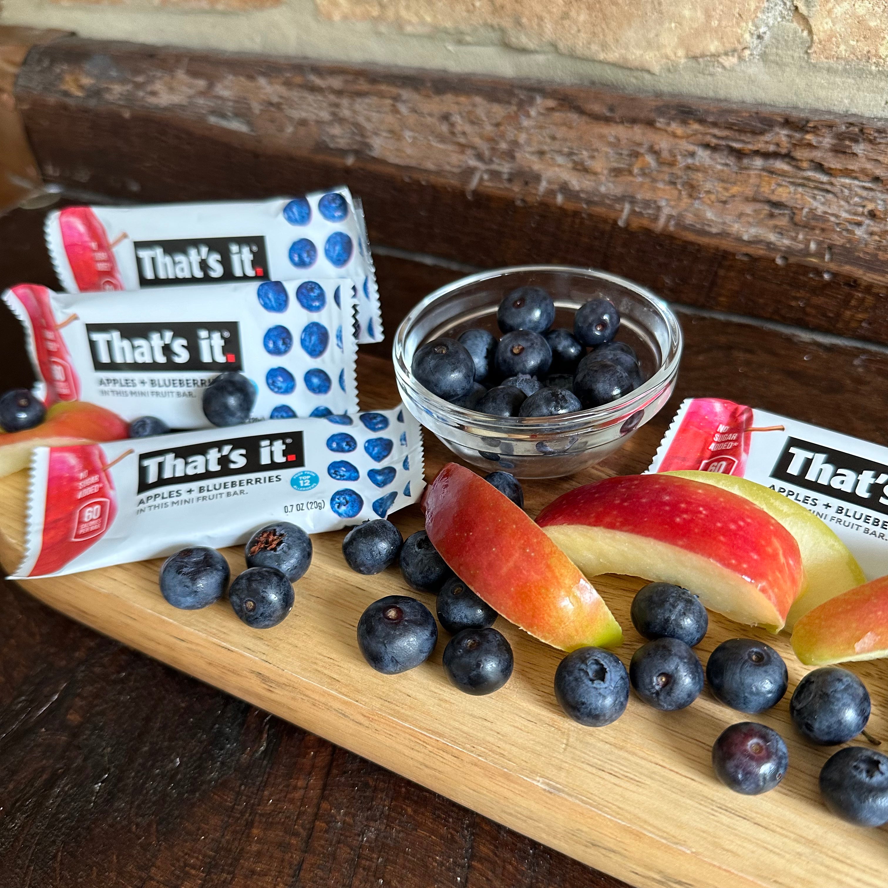 Three That's it Gluten-Free Soft & Chewy Blueberry Mini's Ready-to-Eat Fruit Bars, made with 100% natural non-GMO fruit, are placed on a wooden board beside fresh apple slices and a small bowl of blueberries.