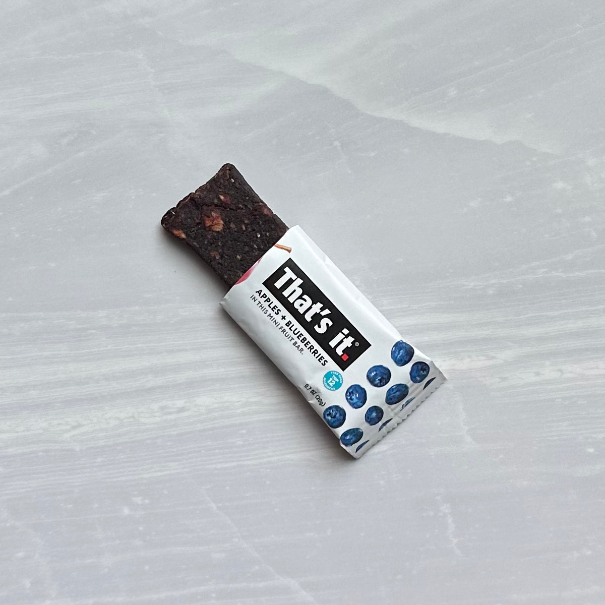 A dark snack bar partially unwrapped, revealing some of its contents. The wrapper reads "That's it" with images of apples and blueberries, indicating it is a "That's it. Gluten-Free Soft & Chewy Blueberry Mini's Ready-to-Eat Fruit Bar, 0.7 oz - 1 Count," made from 100% natural non-GMO fruit.