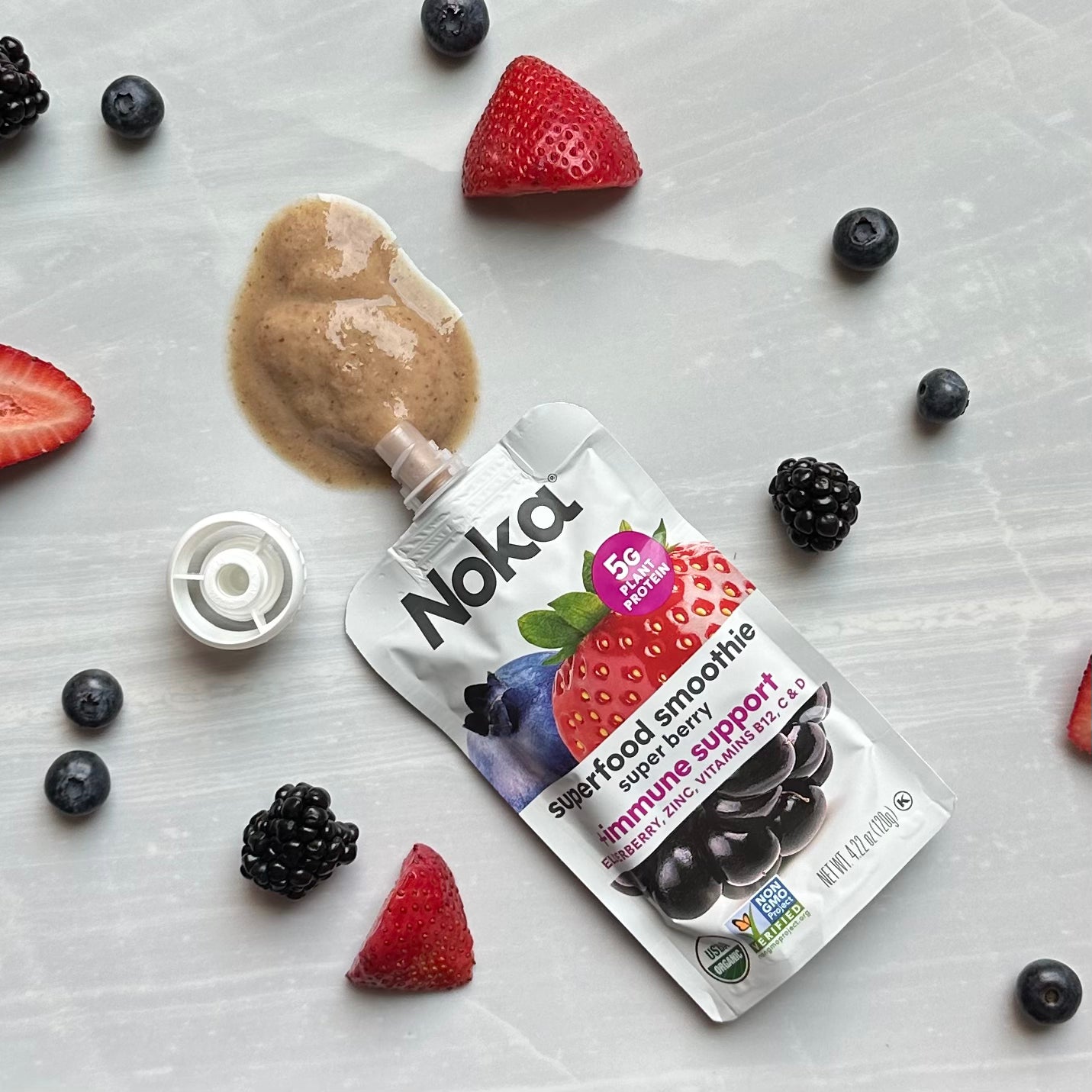 A Noka Organic Super Berry with Plant Protein pouch, 4.22 oz, lies on a surface with an open cap and some of its immune-boosting contents spilled out. Surrounding the convenient on-the-go Noka pouch are sliced strawberries, blueberries, and blackberries.