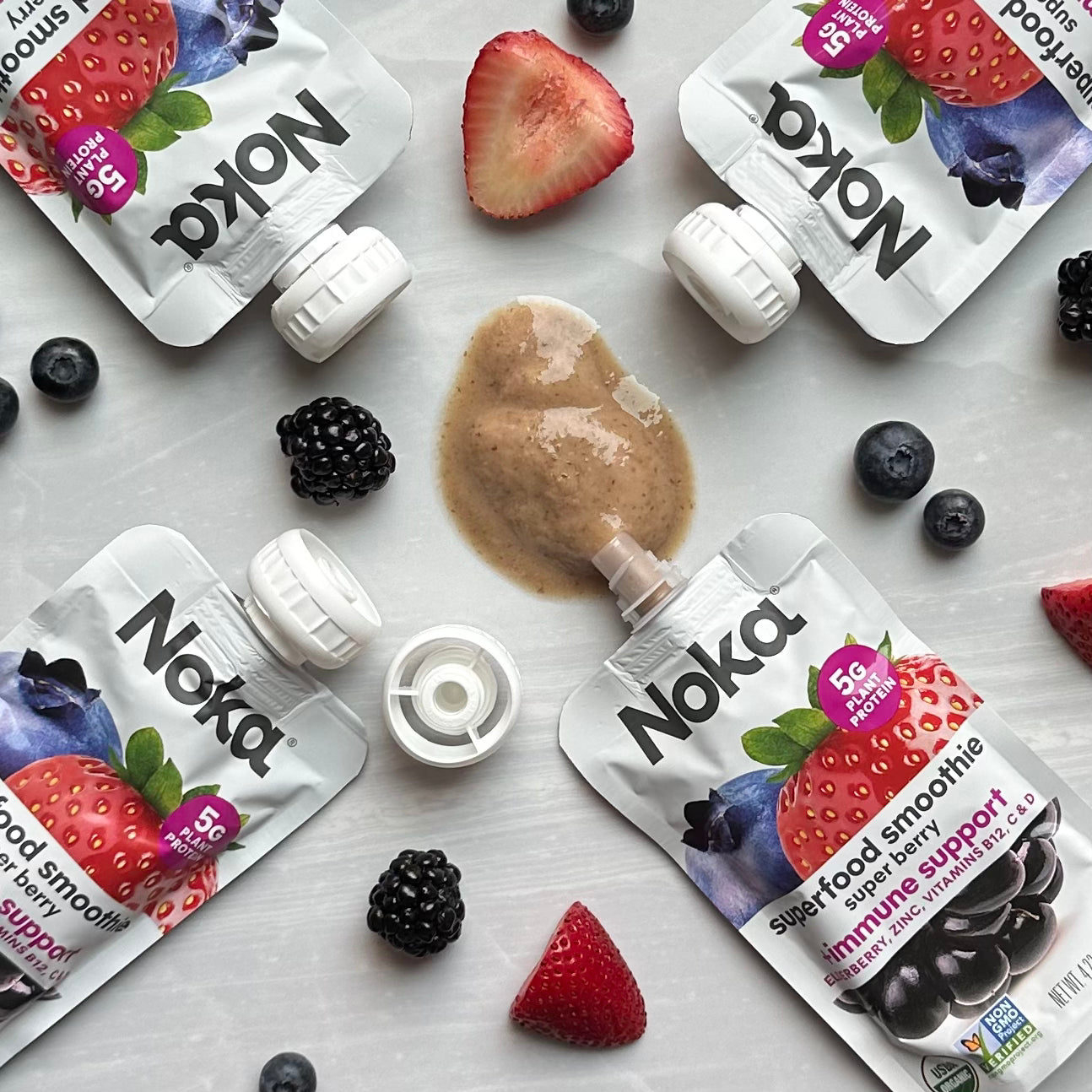 Four Noka Organic Super Berry pouches with Plant Protein lay around scattered blackberries, strawberries, and blueberries on a white surface. One pouch is partially opened with some smoothie spilled—ideal for an on-the-go immune-boosting snack made from organic fruits.