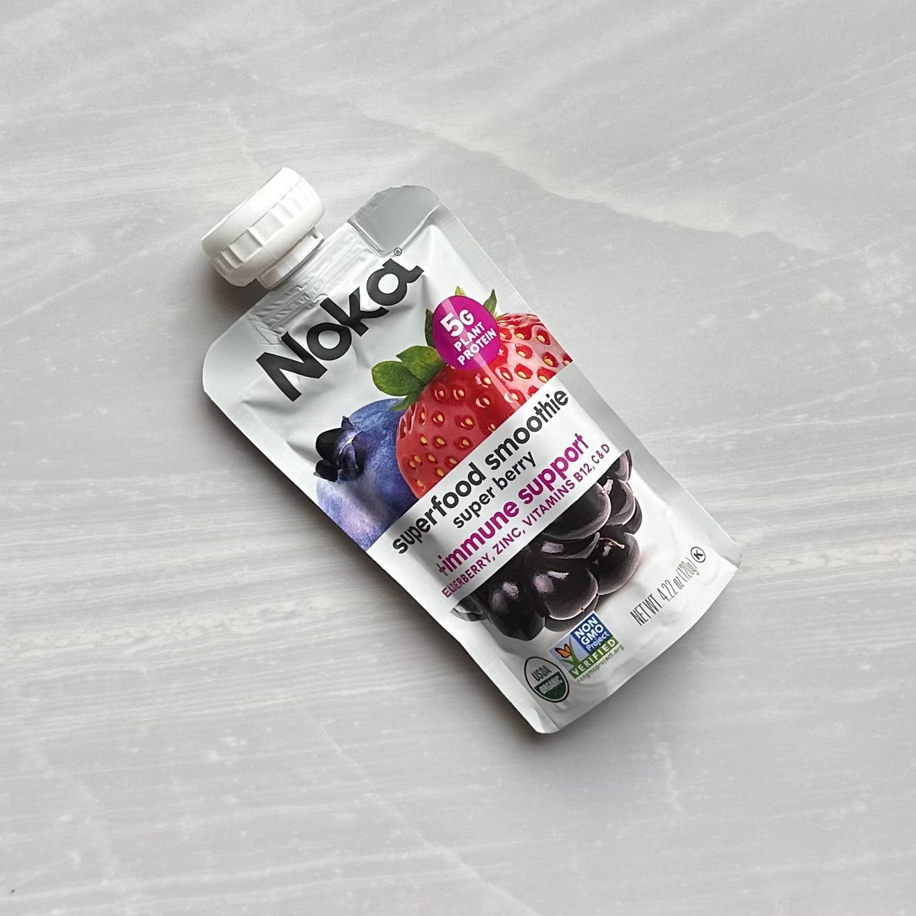 A Noka Organic Super Berry with Plant Protein smoothie pouch, featuring strawberries, blueberries, and other organic fruits on the package. Labeled “Immune Support” with 5g of protein, it's perfect for on-the-go nourishment. Set against a light marble surface, it’s your immune-boosting companion anywhere.