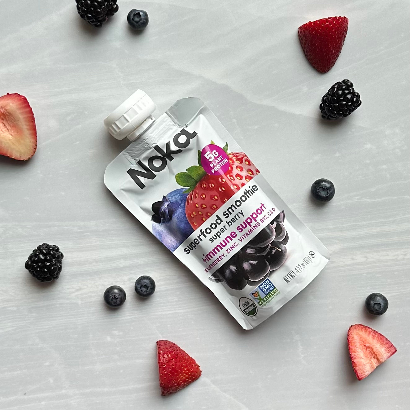 A Noka Organic Super Berry with Plant Protein pouch, packed with organic fruits, sits on a grey background surrounded by scattered blackberries, blueberries, and halved strawberries - an immune-boosting snack perfect for on-the-go.