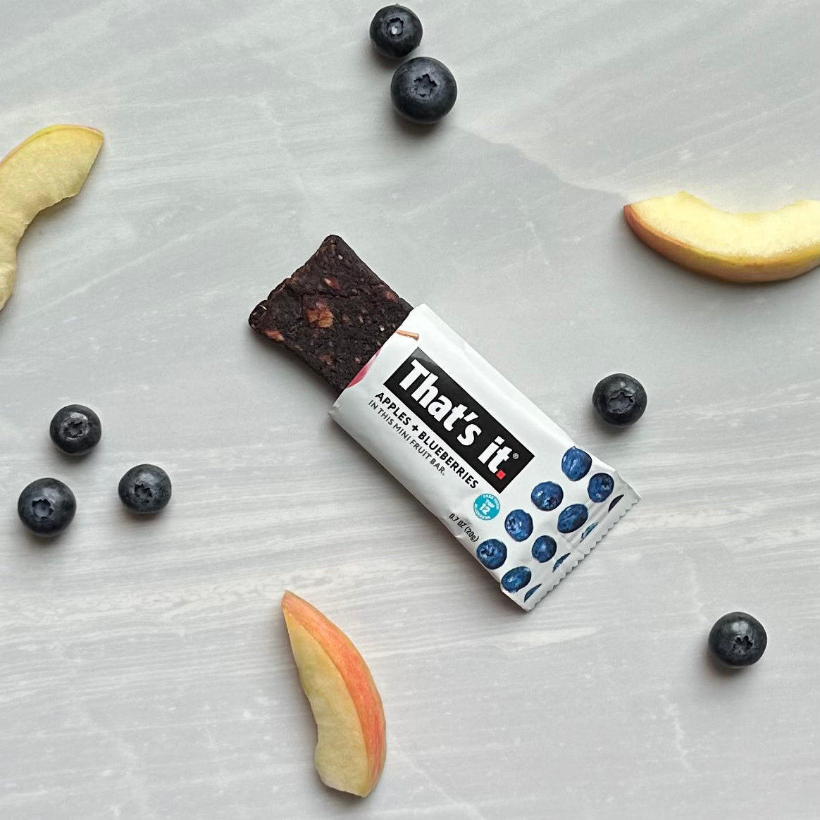 An opened That's it. Gluten-Free Soft & Chewy Blueberry Mini's Ready-to-Eat Fruit Bar, a snack made from 100% natural non-GMO fruit, is surrounded by fresh blueberries and apple slices on a gray surface. This vegan, gluten-free, dairy-free treat is perfect for a healthy snacking option.