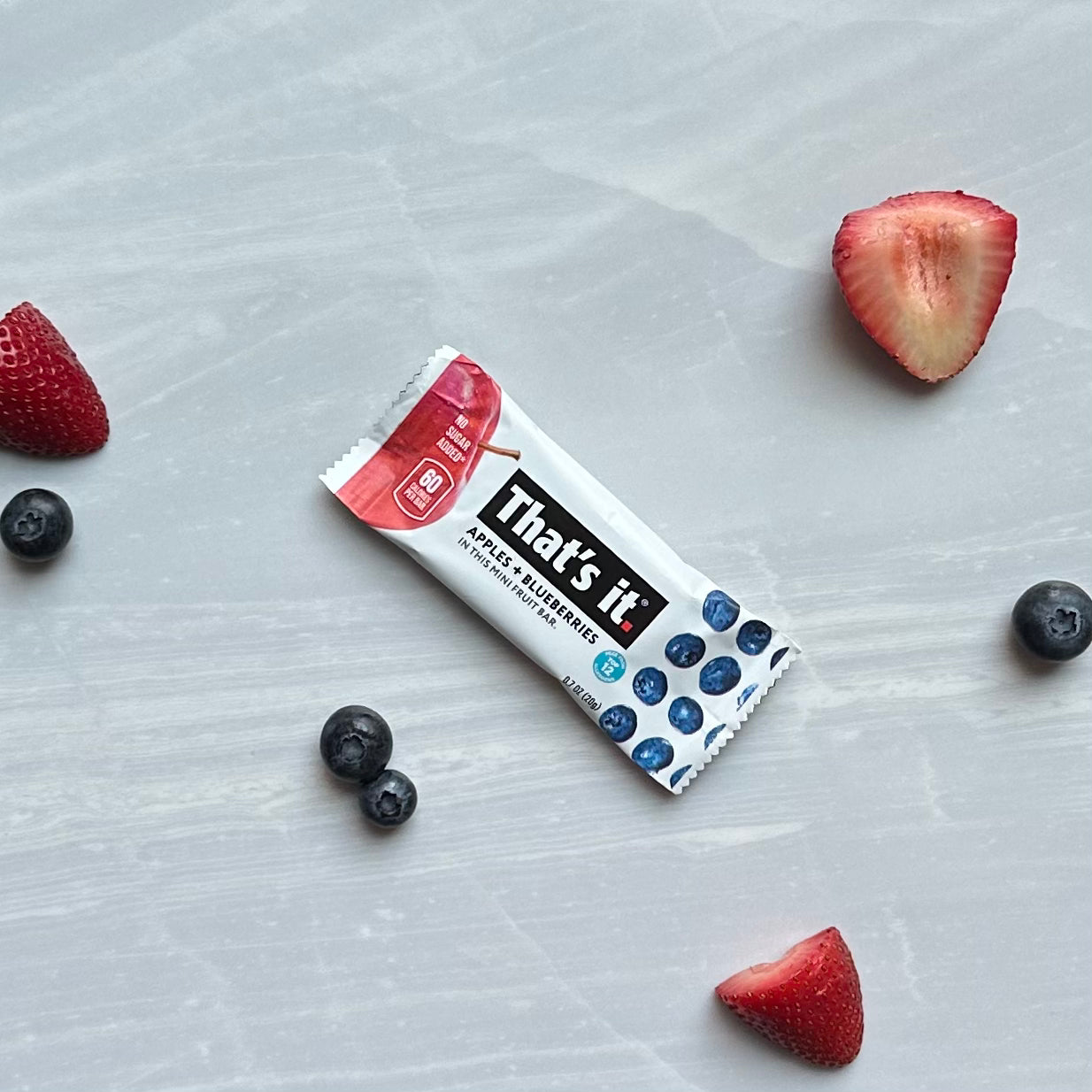 A "That's it. Gluten-Free Soft & Chewy Blueberry Mini's Ready-to-Eat Fruit Bar," an all-natural fruit snack bar, is placed on a gray marble surface, surrounded by scattered strawberries and blueberries.