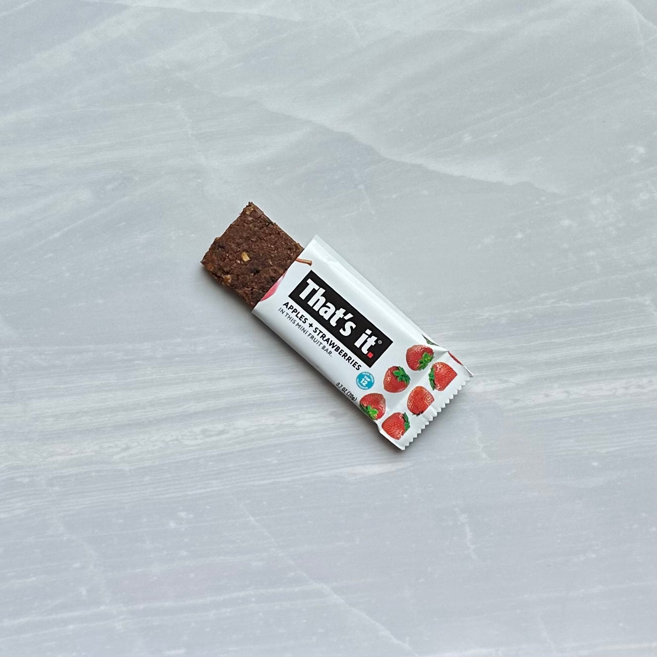 A partially unwrapped fruit bar lies on a gray surface. The packaging features strawberries and the text "That's it." These That's it Strawberry Mini Fruit Bars are a delicious gluten-free, plant-based snack.