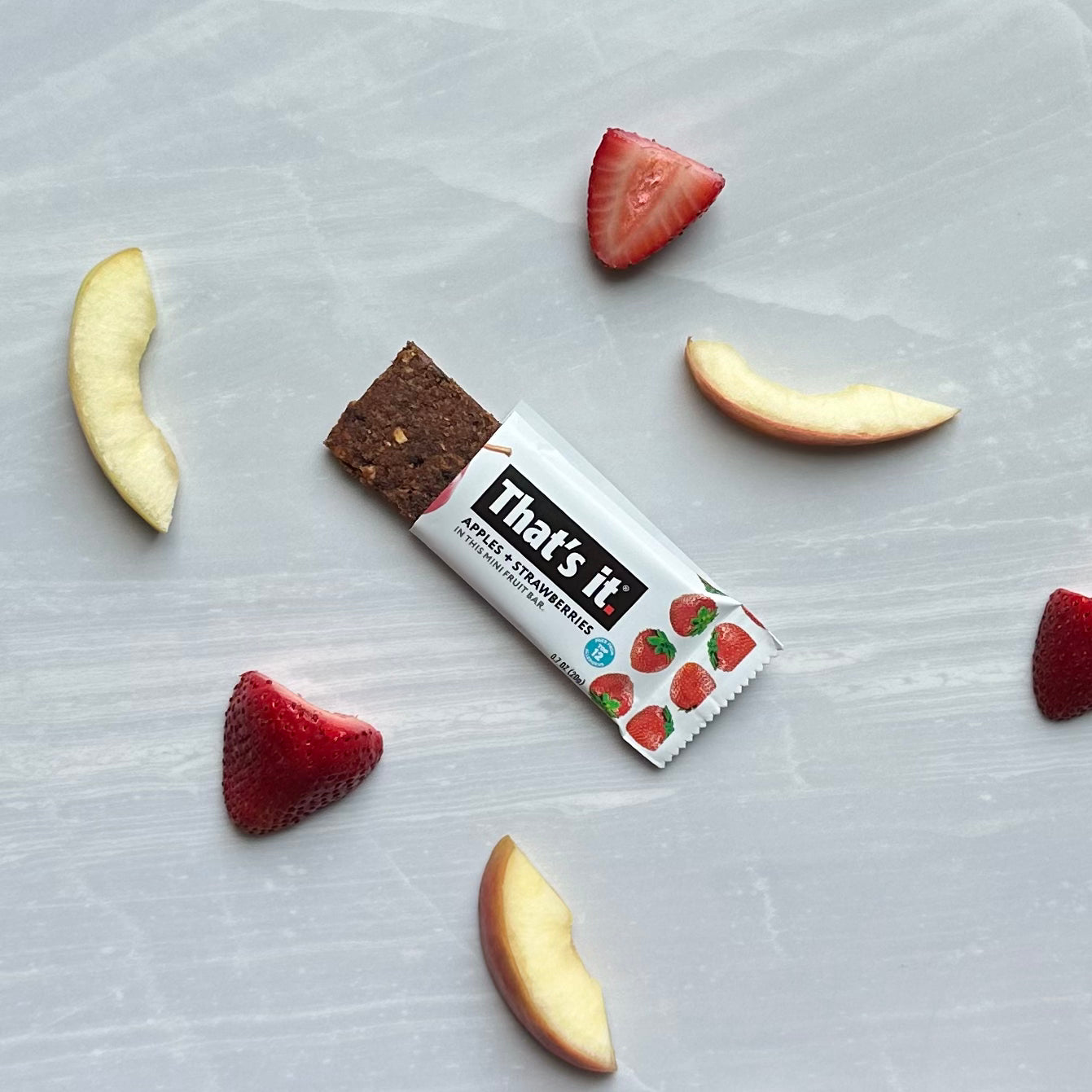 A fruit bar labeled "That's it Strawberry Mini Fruit Bars" is placed on a light gray surface, surrounded by slices of apple and strawberries. This gluten-free, plant-based snack from That's it is perfect for those craving a healthy treat.