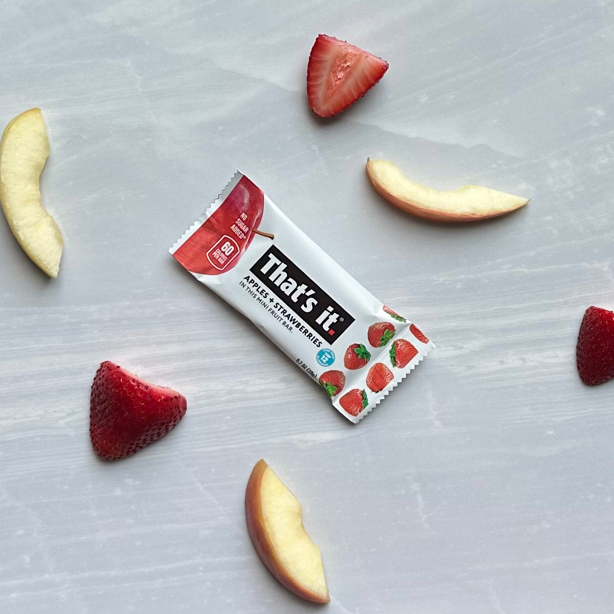 A gluten-free, plant-based snack bar labeled “That's it Strawberry Mini Fruit Bars” lies on a gray surface surrounded by strawberry and apple slices.