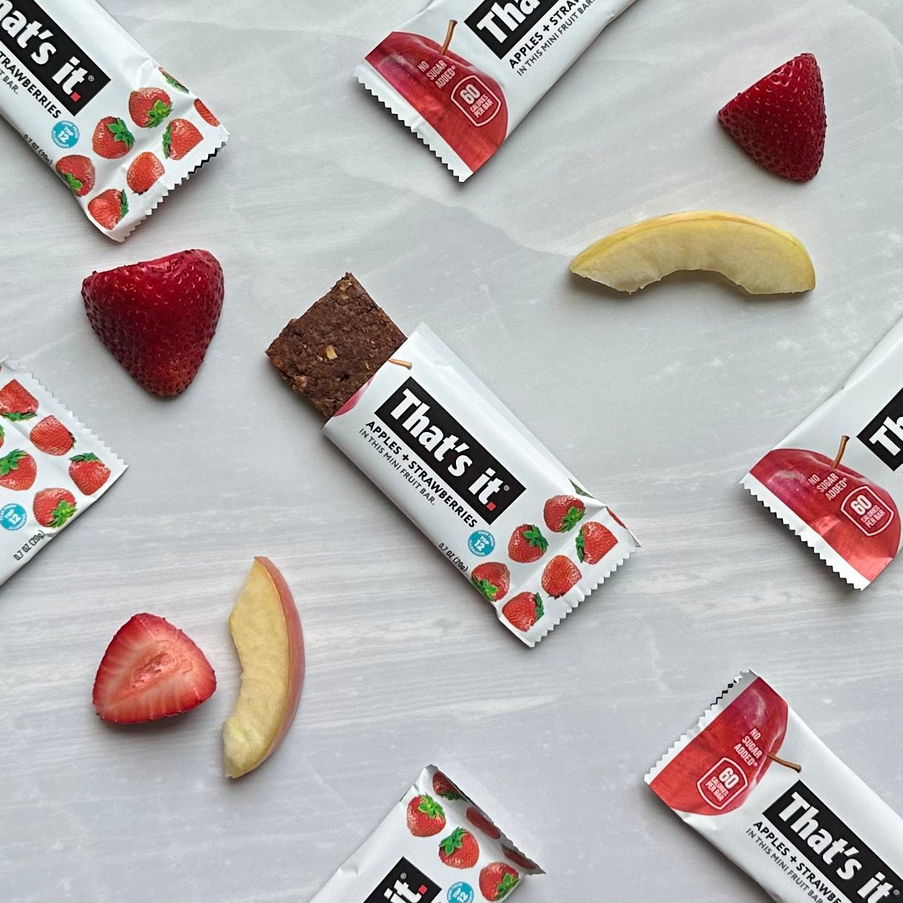 Scattered fruit bars, halved strawberries, and apple slices are arranged on a light surface. The packaging reads "That's it" with images of strawberries and apples. These That's it Strawberry Mini Fruit Bars are a delightful plant-based snack that’s also gluten-free.