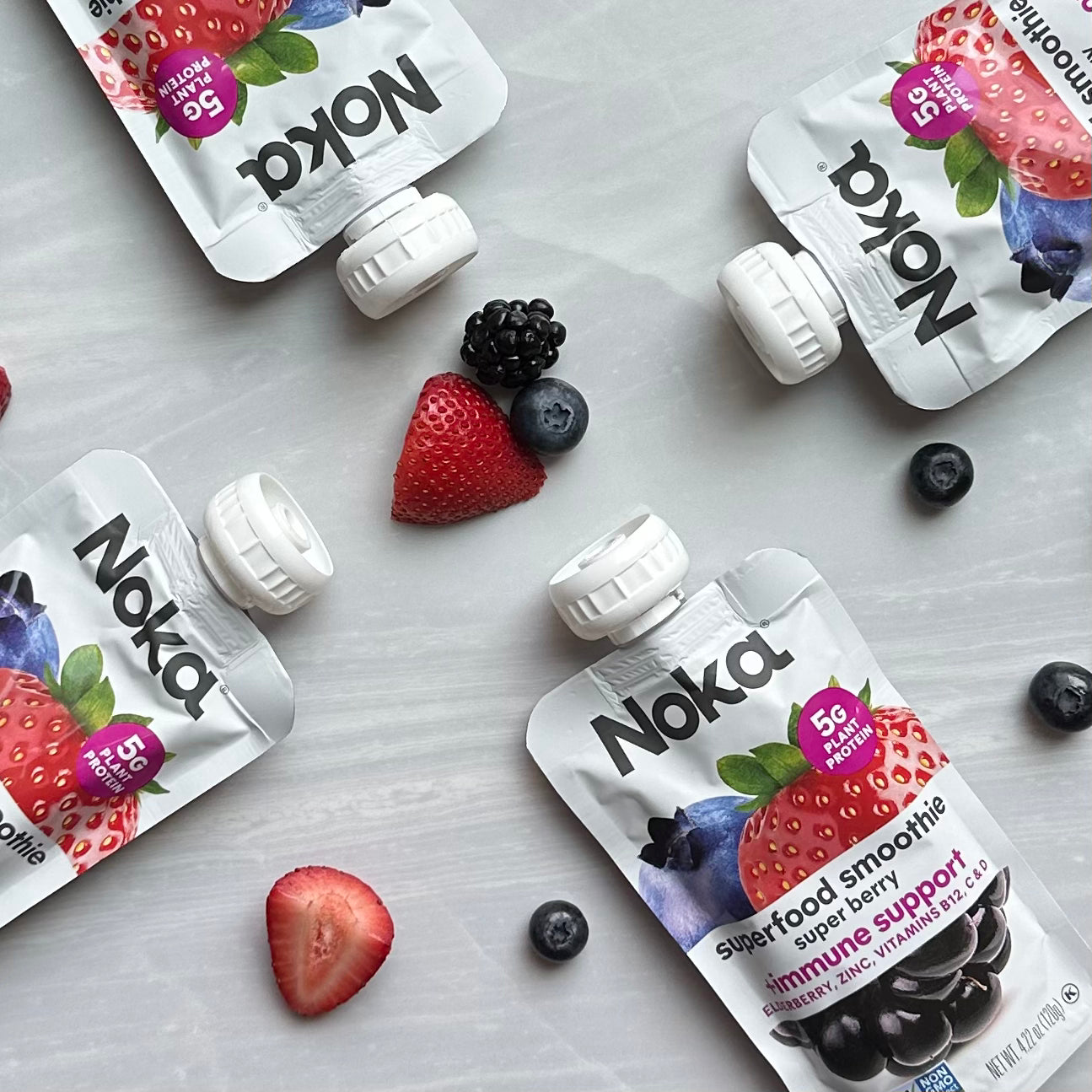 Noka Organic Super Berry with Plant Protein, 4.22 oz, pouches arranged on a light surface with scattered organic fruits like blueberries, blackberries, and a halved strawberry—perfect for an on-the-go immune-boosting treat.