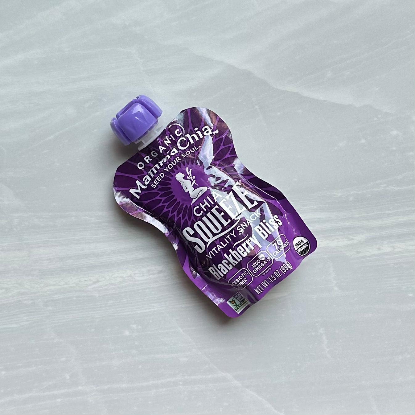 A purple squeezable pouch labeled "Mamma Chia Squeeze Vitality Snack, Blackberry Bliss, 3.5 oz - 1 Count" lies on a white marble surface, boasting organic chia seeds for a boost of omega-3s and natural vitality.