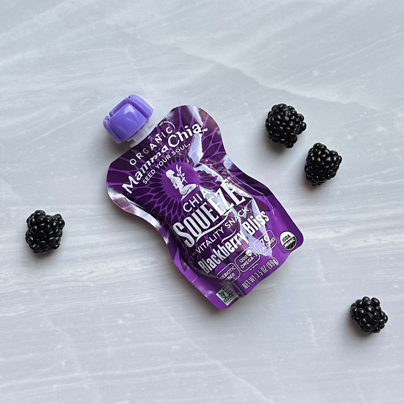 A purple squeeze pouch labeled "Mamma Chia Squeeze Vitality Snack, Blackberry Bliss" with a blackberry bliss flavor, rich in omega-3s and natural vitality, is surrounded by four blackberries on a light gray surface.