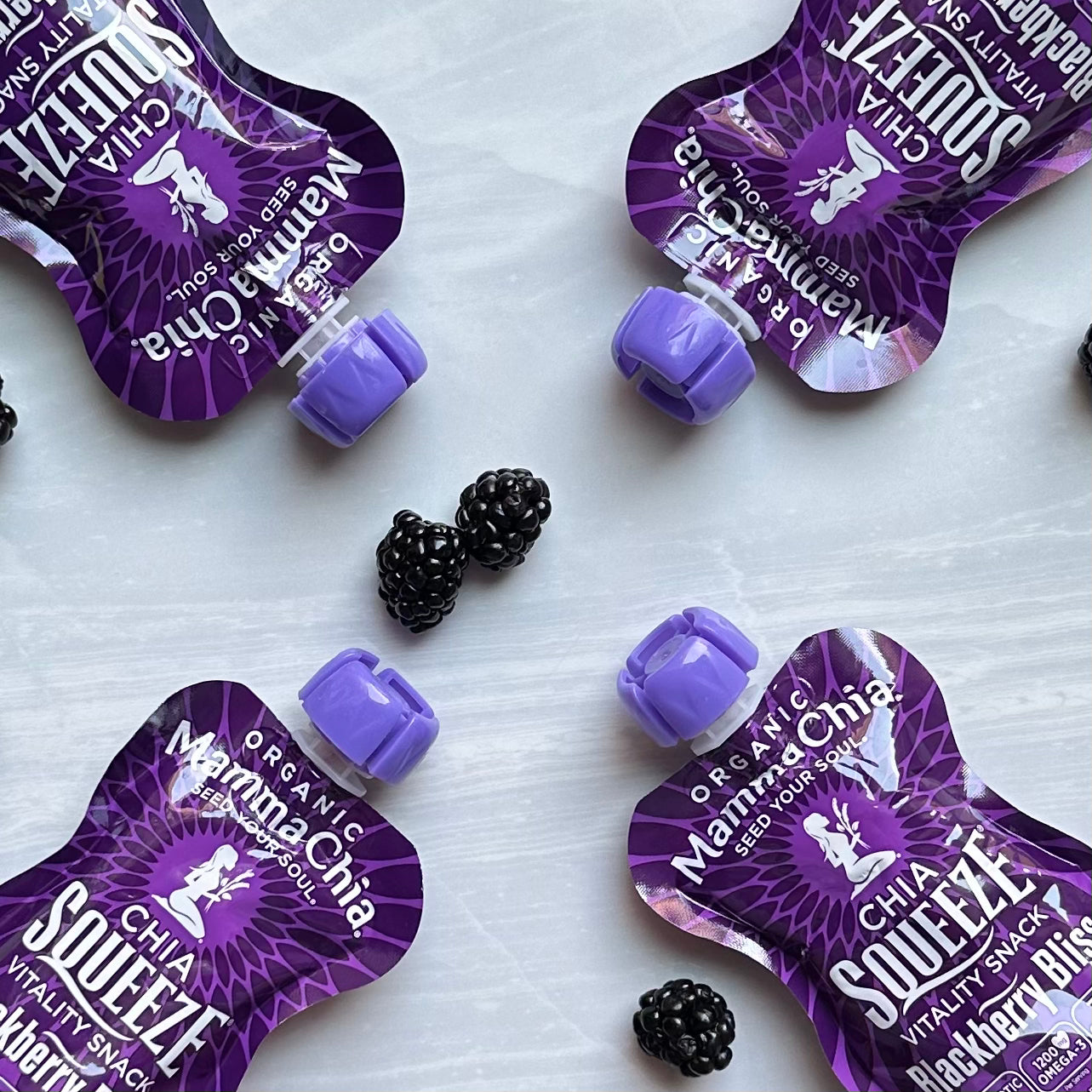 Four packets of the Mamma Chia Squeeze Vitality Snack in Blackberry Bliss flavor, each 3.5 oz and made with organic chia seeds and omega-3s for natural vitality, are placed on a light surface, surrounded by scattered blackberries.