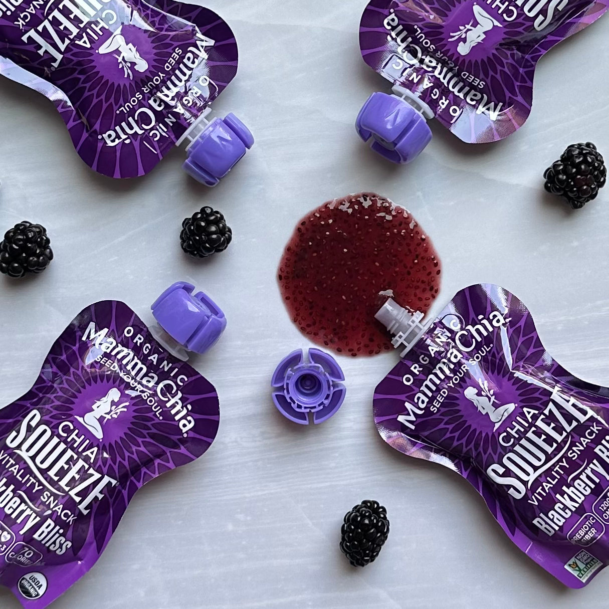 Four Mamma Chia Squeeze Vitality Snack pouches in Blackberry Bliss are arranged in a circle with some blackberries scattered around. One pouch is open, its rich organic chia seed contents spilling into the center of the image. The vibrant display highlights the natural vitality and benefits like omega-3s.