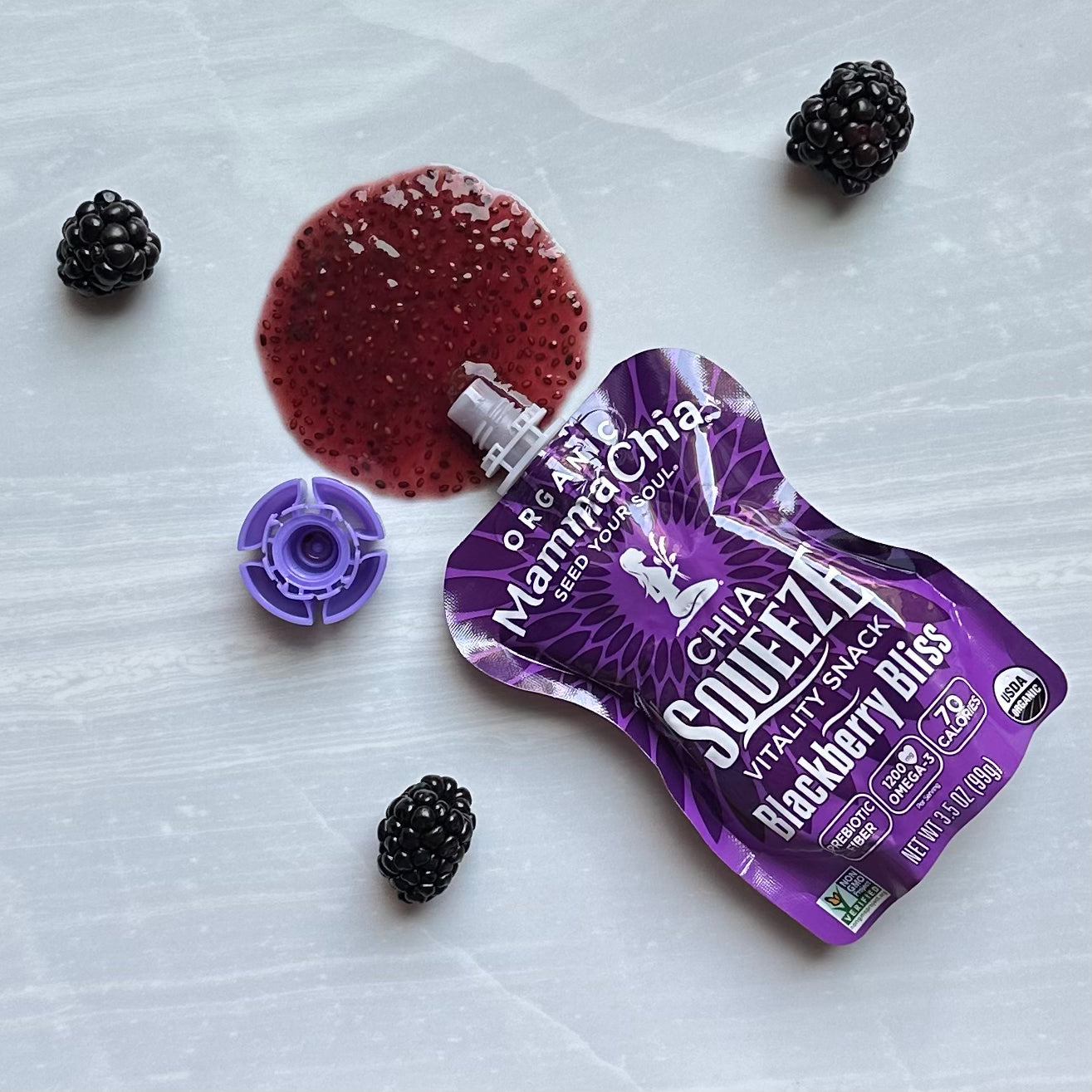 A spilled pack of Mamma Chia Squeeze Vitality Snack, Blackberry Bliss, 3.5 oz - 1 Count with some blackberries around it on a white surface. The pack, rich in organic chia seeds and omega-3s, is opened and the contents are partially spilled, showcasing its promise of natural vitality from the trusted brand, Mamma Chia.