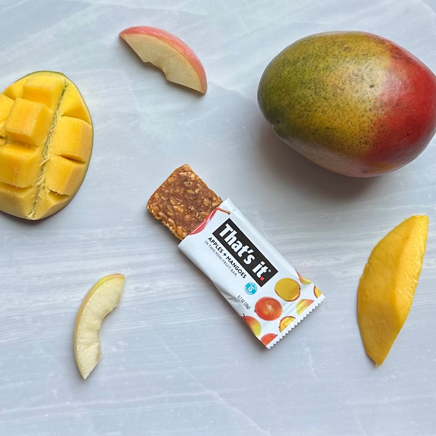 A partially unwrapped "That's it. Gluten-Free Soft & Chewy Mango Mini Fruit Bar, 0.7 oz - 1 Count" lies on a surface surrounded by slices and a whole mango, along with some sliced apple pieces. This all-natural fruit snack from That's it is vegan, gluten-free, and contains no added sugars.