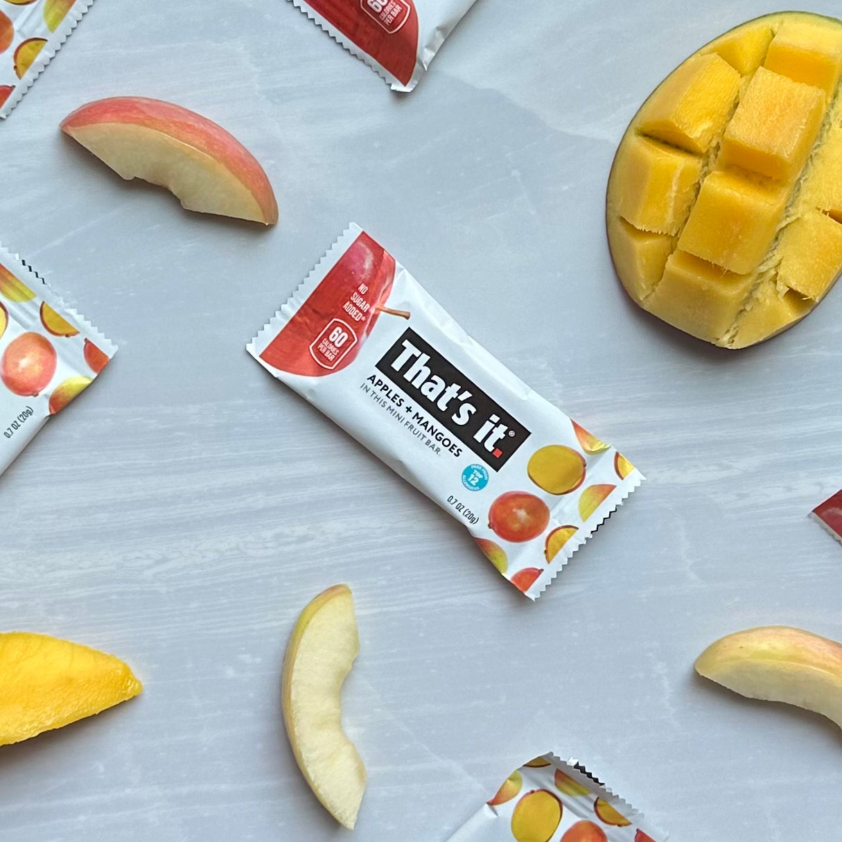 A fruit snack bar labeled "That's it. Gluten-Free Soft & Chewy Mango Mini Fruit Bar, 0.7 oz - 1 Count" is surrounded by slices of apple and a partially diced mango on a light-colored surface, showcasing an all-natural fruit snack that's vegan, gluten-free, and contains no added sugars.