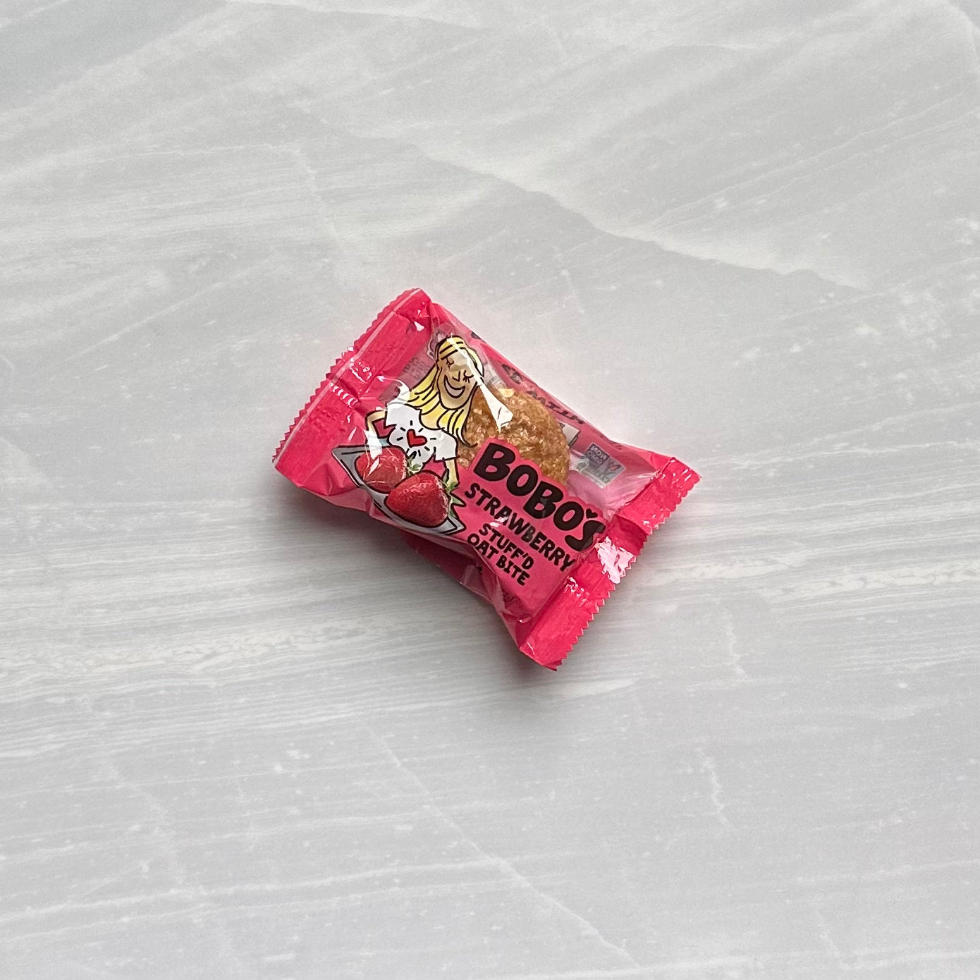 A light gray marbled surface holds a Bobo's Strawberry Stuff'd Oat Bite, wrapped in a red package and proudly labeled vegan and gluten-free.