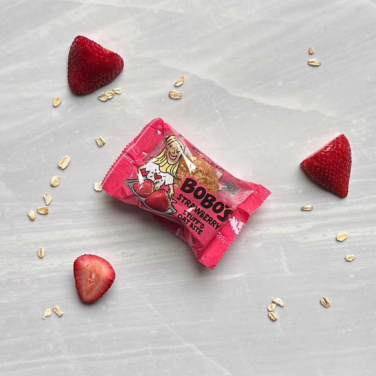 A Bobo's Strawberry Stuff'd Oat Bite in pink packaging is placed on a light gray surface, surrounded by three strawberry halves and scattered oat flakes, showcasing its gluten-free, vegan ingredients.