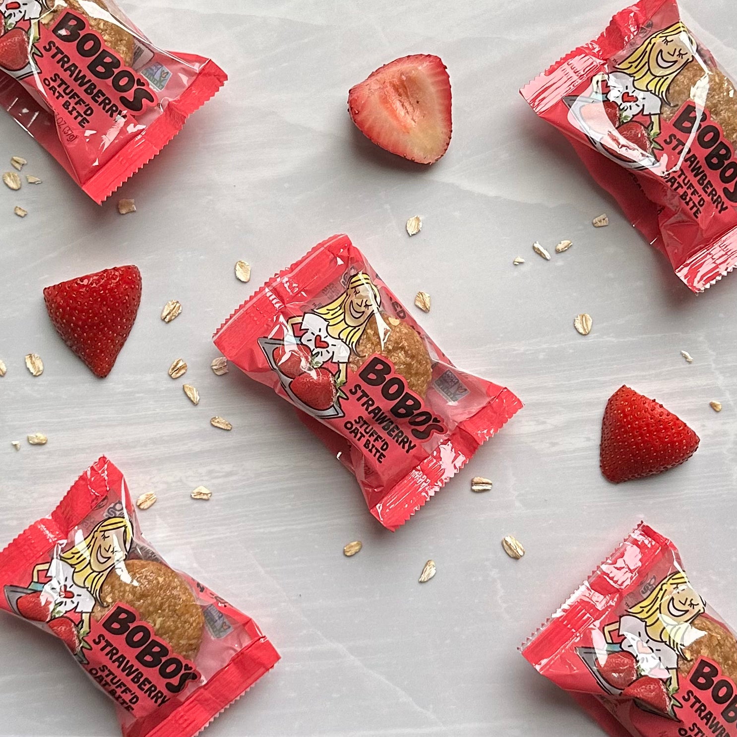 Five individual packs of Bobo's Strawberry Stuff'd Oat Bites, a delightful gluten-free and vegan snack, are arranged on a light surface along with fresh strawberry halves and scattered oats.