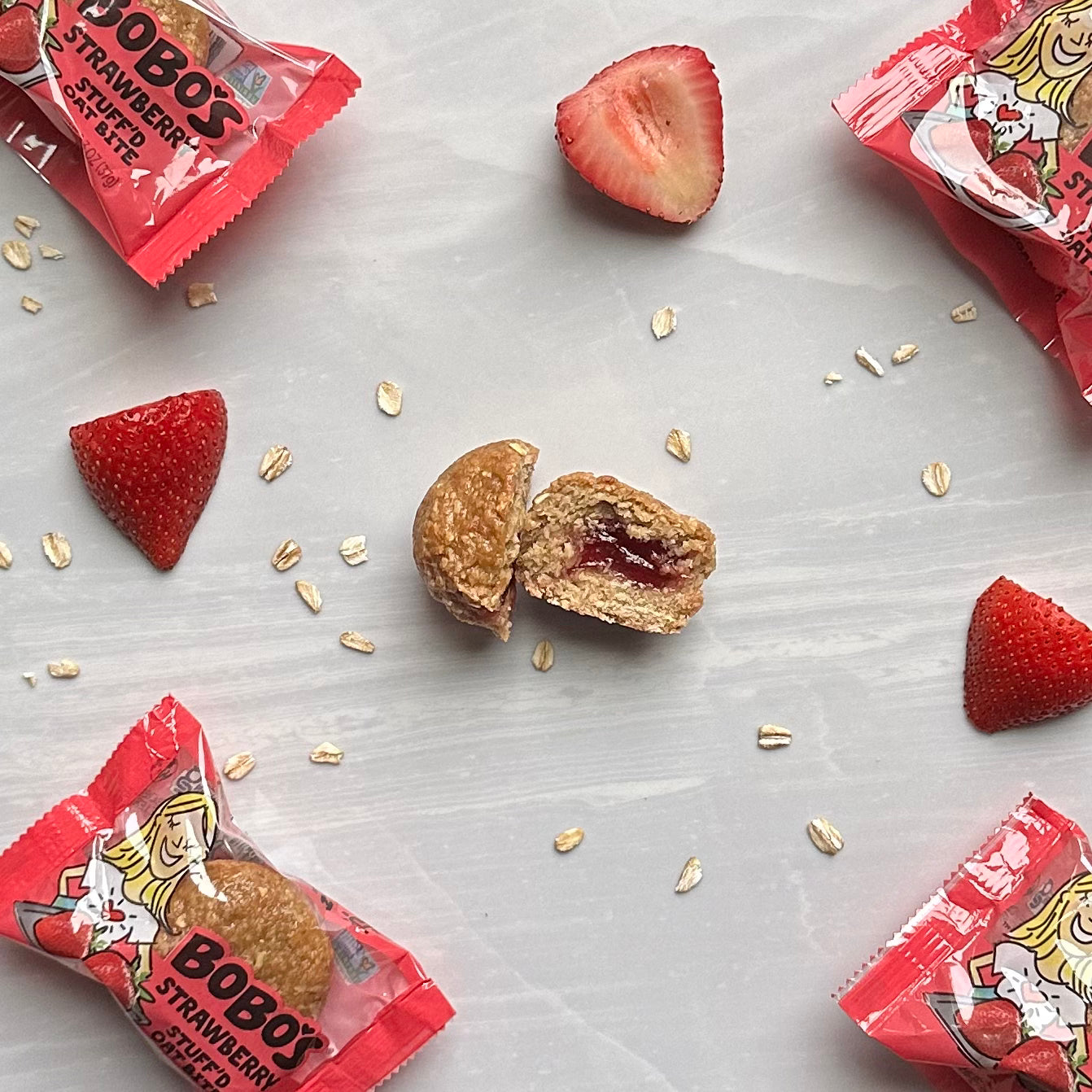 A halved Bobo's Strawberry Stuff'd Oat Bite is surrounded by strawberry pieces, oats, and wrapped Bobo's Oat Bites on a light-colored surface.