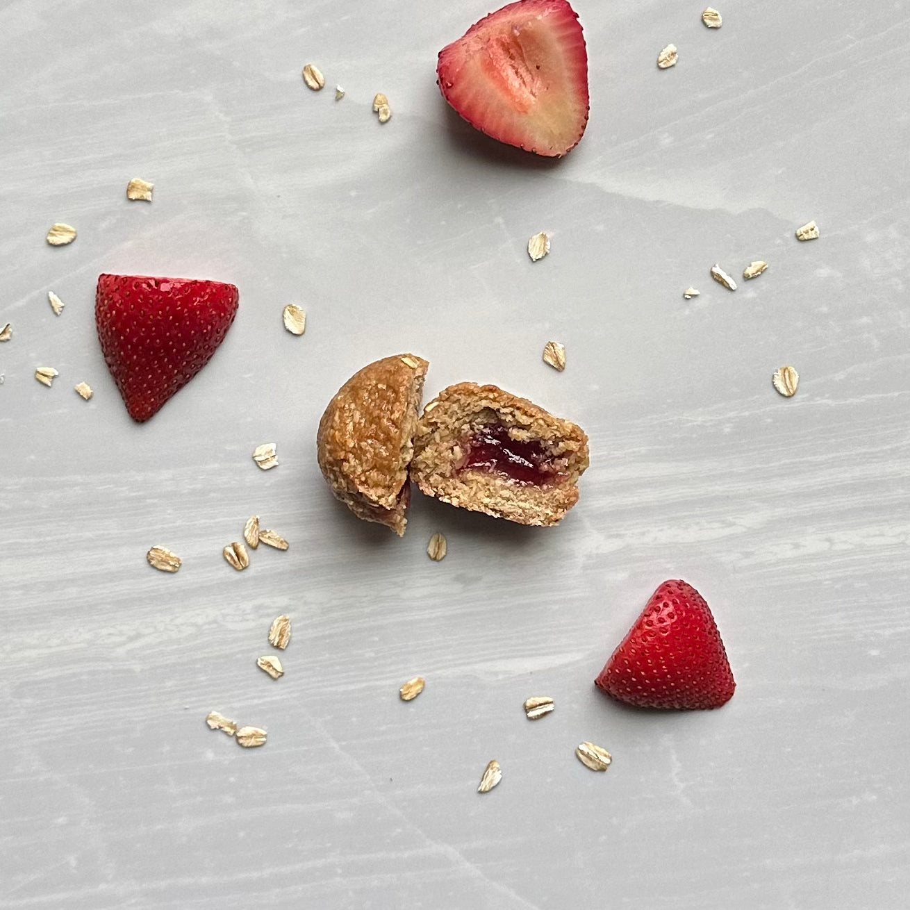 A sliced Bobo's Strawberry Stuff'd Oat Bite - 1.3 oz - 1 Count, filled with strawberry jam, surrounded by strawberry halves and oats scattered on a light gray surface.