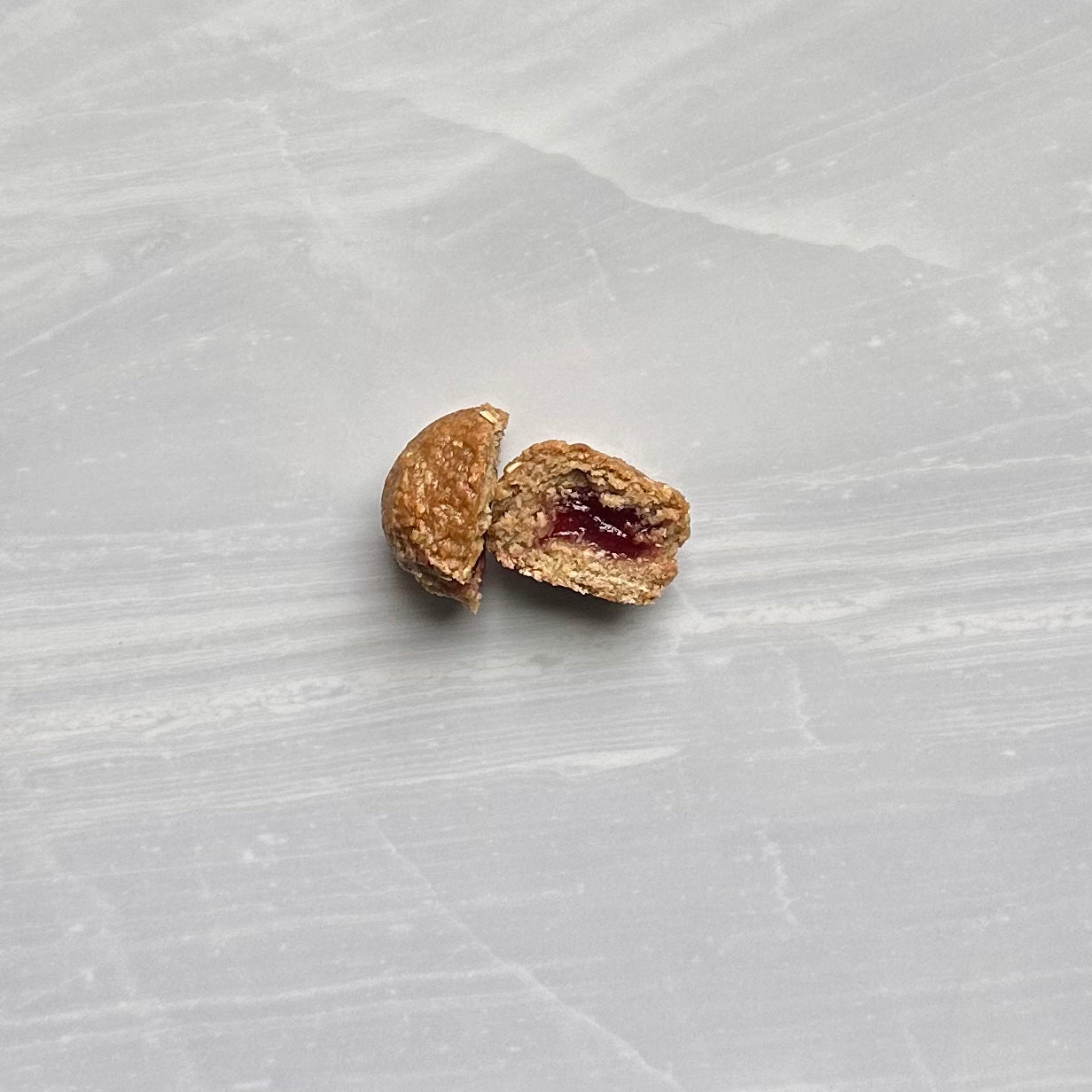 A small, round Bobo's Strawberry Stuff'd Oat Bite, broken in half to reveal its reddish filling, sits on a light grey, textured surface.