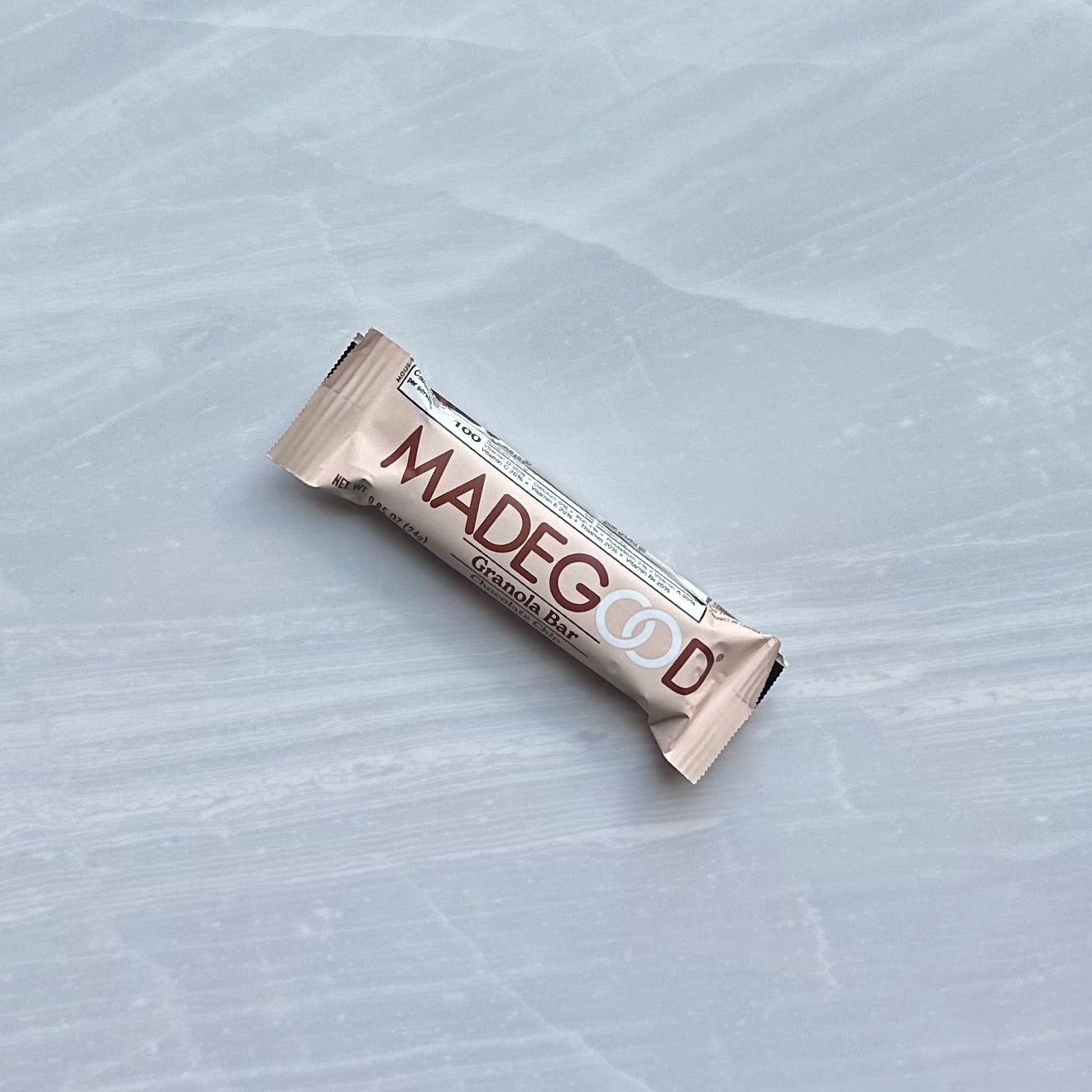 A sealed, beige wrapper of a MadeGood Mixed Berry Granola Bar rests on a smooth, muted gray surface. The text "MADEGOOD" is prominently displayed on the wrapper, hinting at the gluten-free oats and non-GMO ingredients inside this vegan snack.