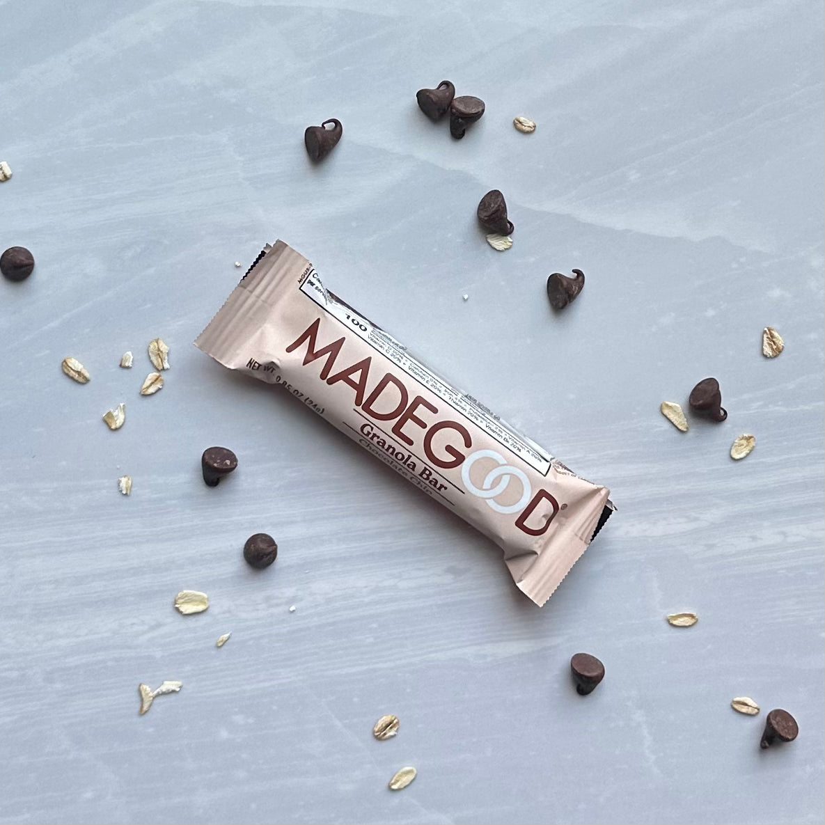 A MadeGood Mixed Berry Granola Bar lies on a grey surface, surrounded by scattered chocolate chips and gluten-free oats.