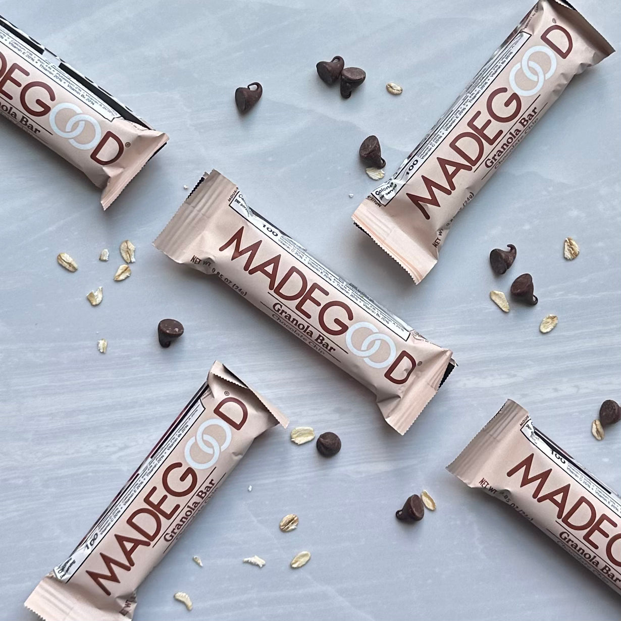 Five MadeGood Mixed Berry Granola Bars, with three unwrapped and two wrapped, are displayed on a light surface adorned with scattered gluten-free oats and chocolate chips. These vegan snacks from MadeGood are also non-GMO, making them a wholesome choice for any time of day.