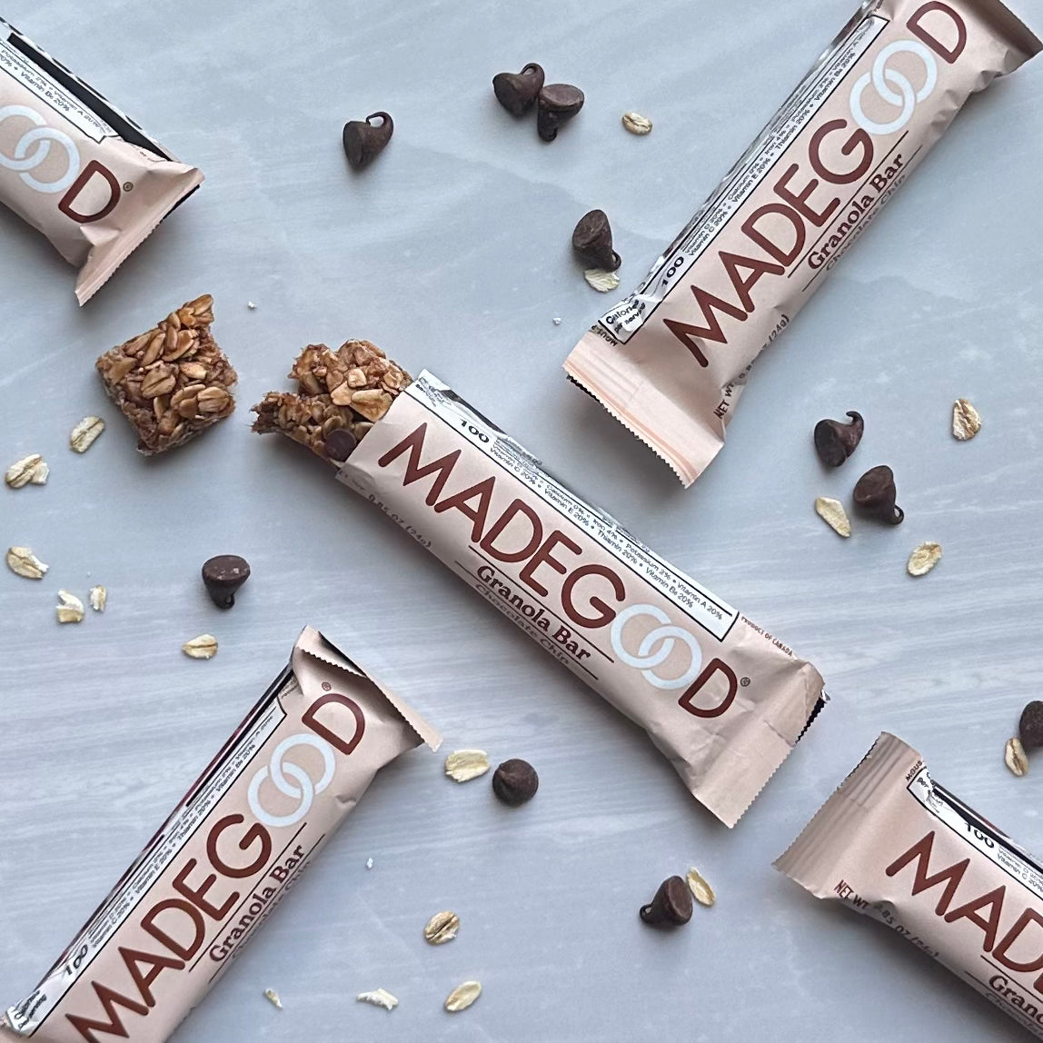 Four MadeGood Mixed Berry Granola Bars, partially unwrapped, are displayed on a light surface surrounded by scattered chocolate chips and non-GMO, gluten-free oats.