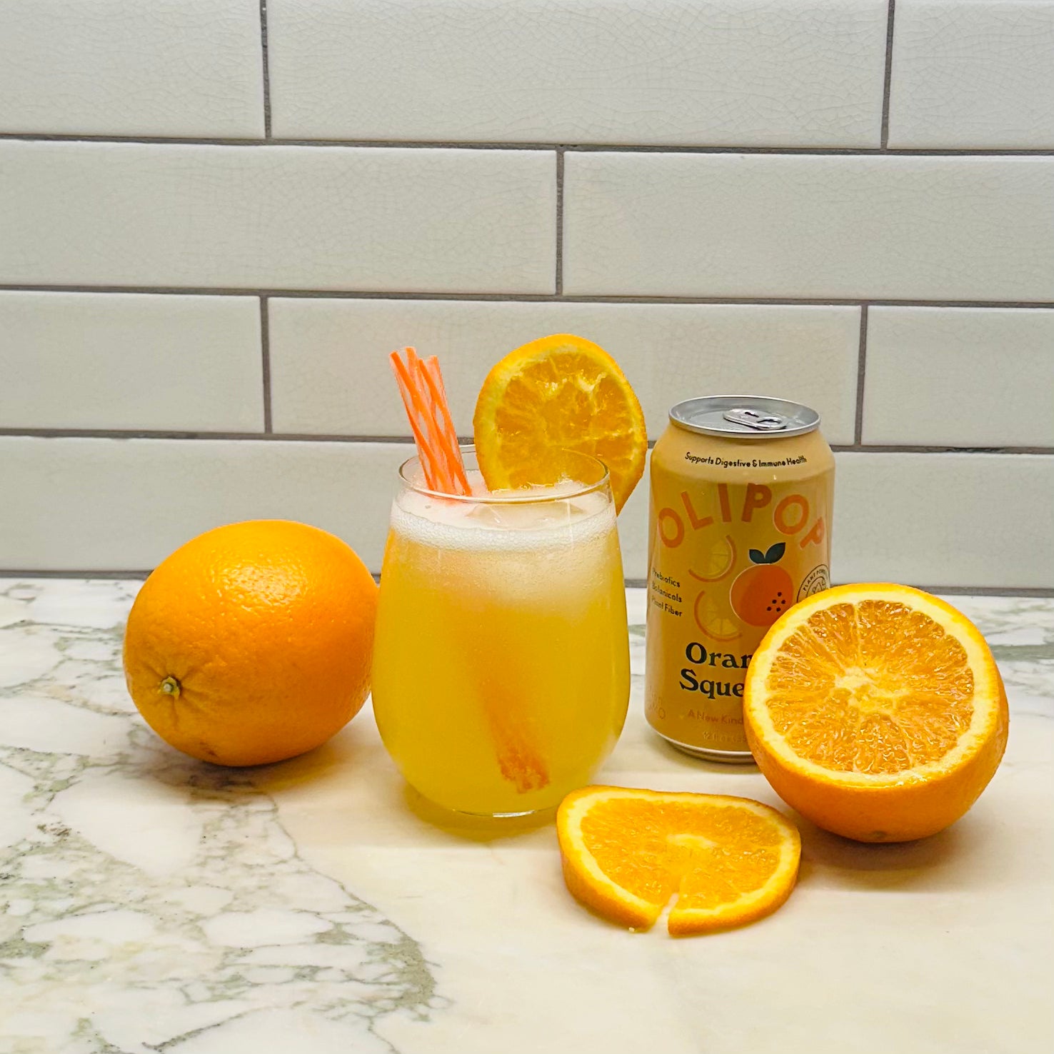 Glass of orange soda with straws, surrounded by whole and sliced oranges, next to a can of Olipop Tropical Punch, a plant-based beverage that promotes gut health with prebiotics, placed on a marble countertop with a white tiled wall in the background.