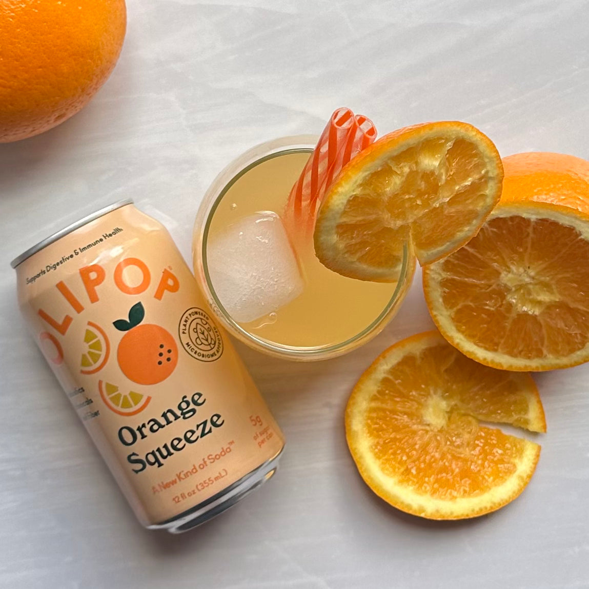 A can of OLIPOP Tropical Punch soda, a plant-based beverage promoting gut health with prebiotics, sits beside a glass with ice and a striped straw, while halved oranges are arranged on a light surface.