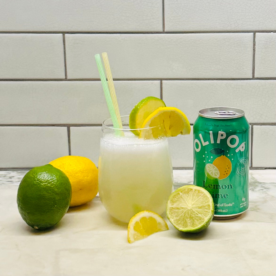 A glass of lemon-lime soda with two straws and garnished with lemon and lime slices, placed beside a can of OLIPOP Prebiotic Soda, Lemon Lime - 12 fl oz - 1 Count by Olipop, known for its digestive health benefits with prebiotics, a whole lemon, a whole lime, and a cut lime on a tiled counter.