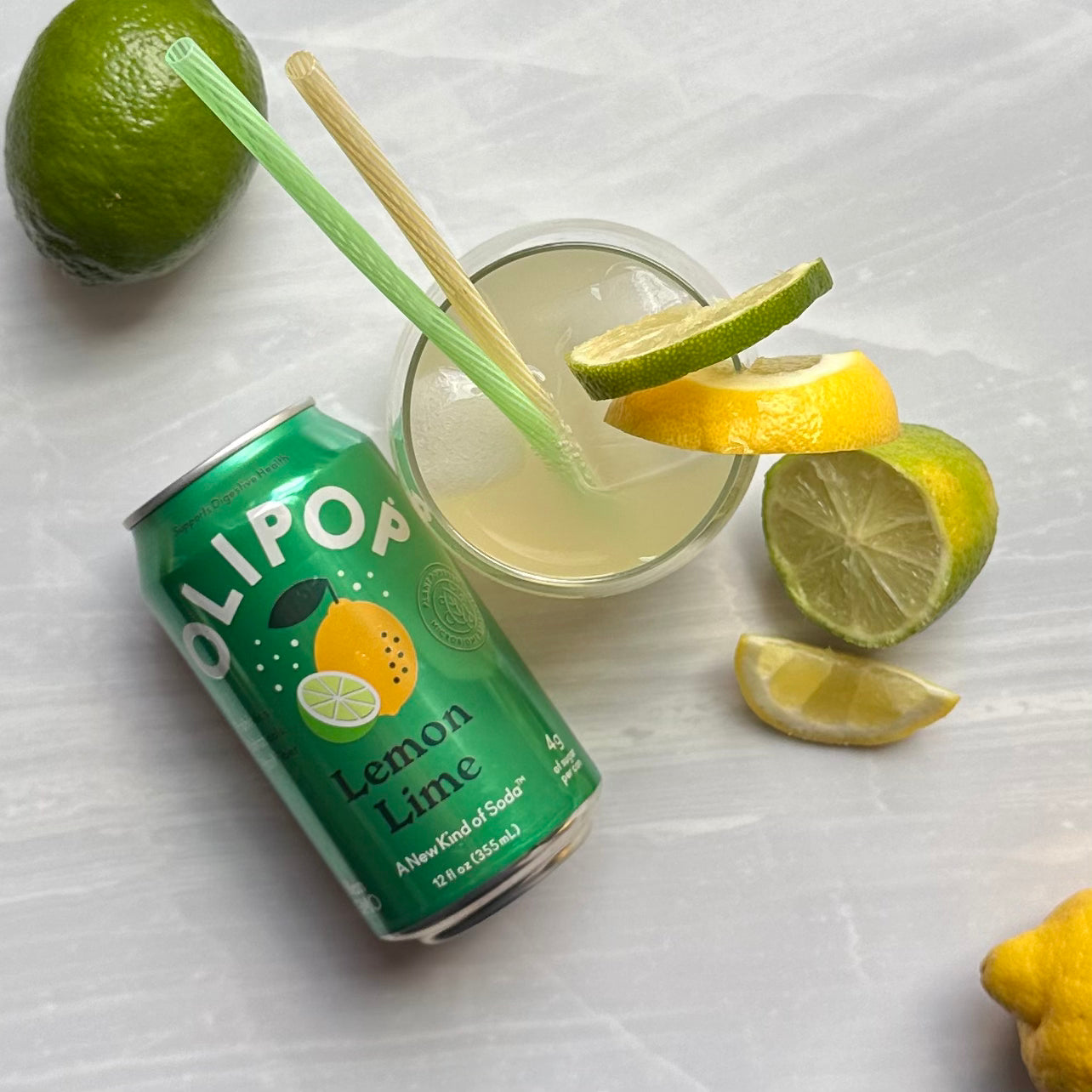 A can of OLIPOP Prebiotic Soda, Lemon Lime flavor (12 fl oz), sits next to a glass of lemon-lime drink with two straws, surrounded by a whole lime, a sliced lime, and lemon wedges on a white surface. This vegan-friendly beverage from Olipop is rich in prebiotics for digestive health, offering both flavor and benefits.