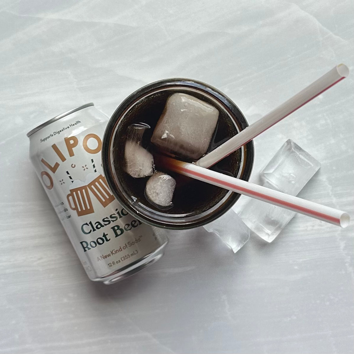 Next to a glass filled with ice and root beer, featuring two straws and two ice cubes placed beside the glass, sits a can of OLIPOP Prebiotic Soda, Classic Root Beer - 12 fl oz - 1 Count. This low-sugar beverage from Olipop is crafted with prebiotics for your digestive health.