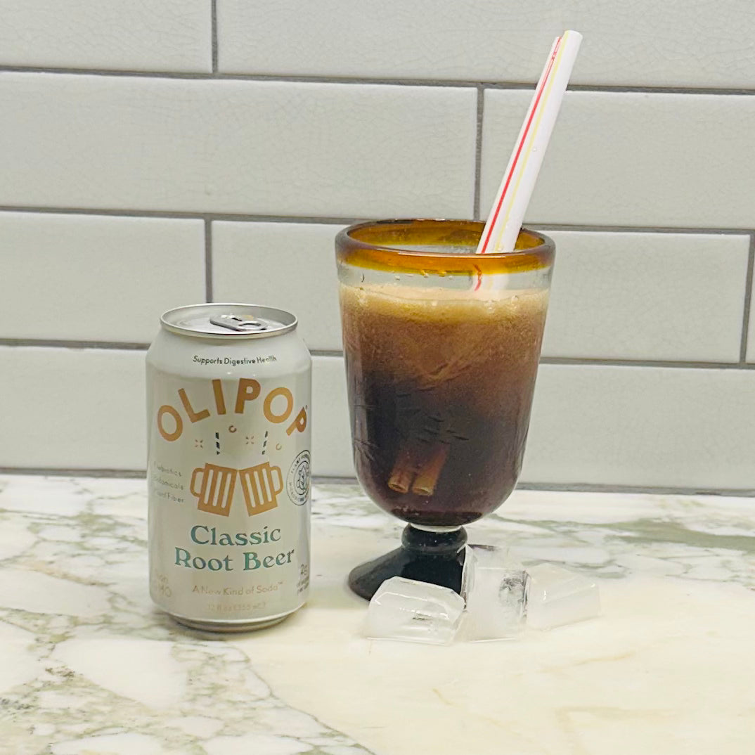 An Olipop Prebiotic Soda, Classic Root Beer - 12 fl oz can, celebrated for its prebiotics and low sugar content, sits next to a glass filled with root beer and ice, adorned with a red and white striped straw, on a marble countertop against a tiled wall.