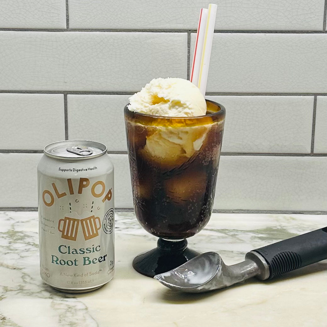 A can of OLIPOP Prebiotic Soda, Classic Root Beer - 12 fl oz - 1 Count, known for its low sugar and prebiotics, sits next to a glass filled with root beer and a scoop of ice cream, with a metal ice cream scooper lying on the counter.