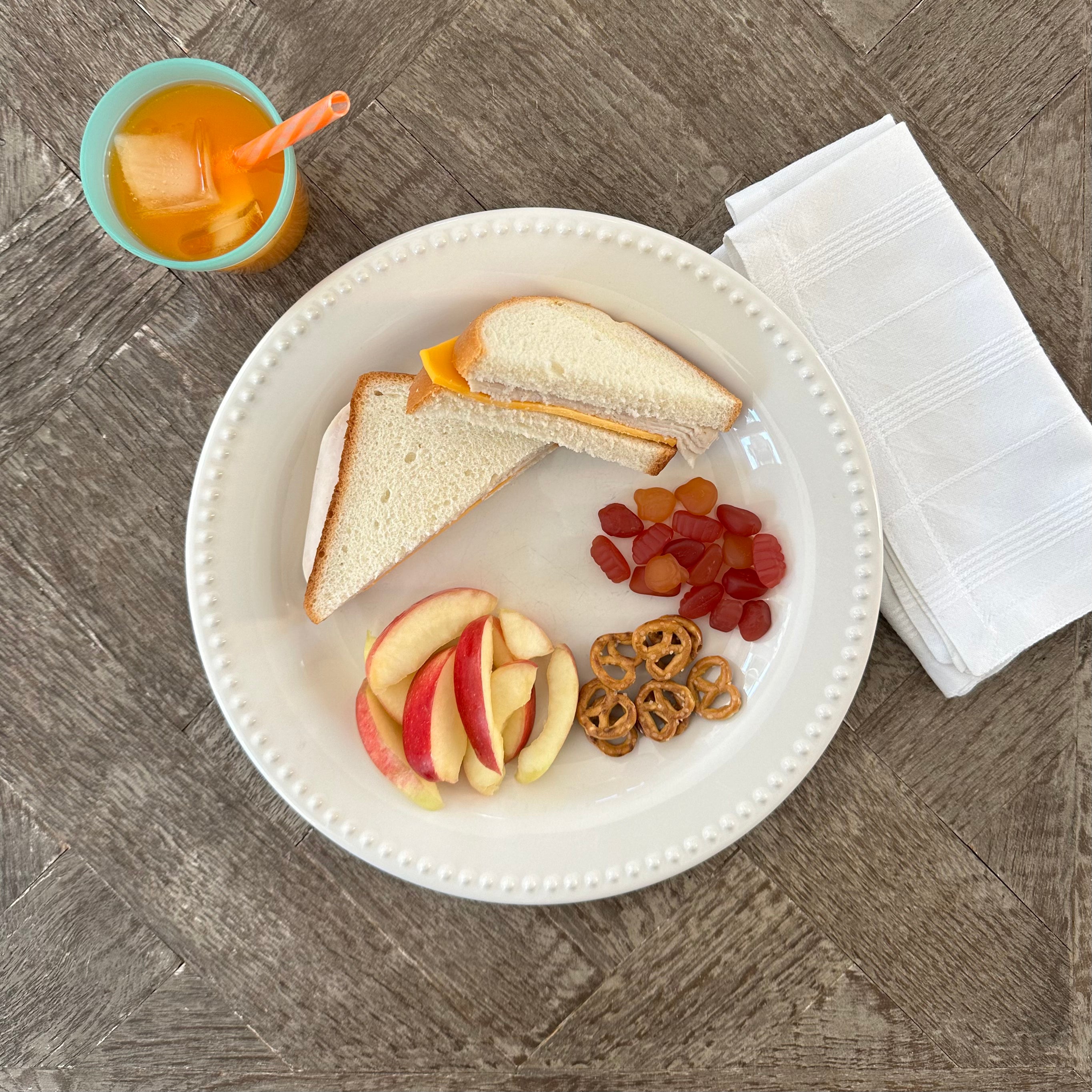 A plate with a Taylor Farms Turkey and Cheddar Sandwich on white bread, served alongside apple slices, gummy candies, and pretzels. A napkin is placed beside the plate. To the left is a glass of orange juice with ice and a straw.