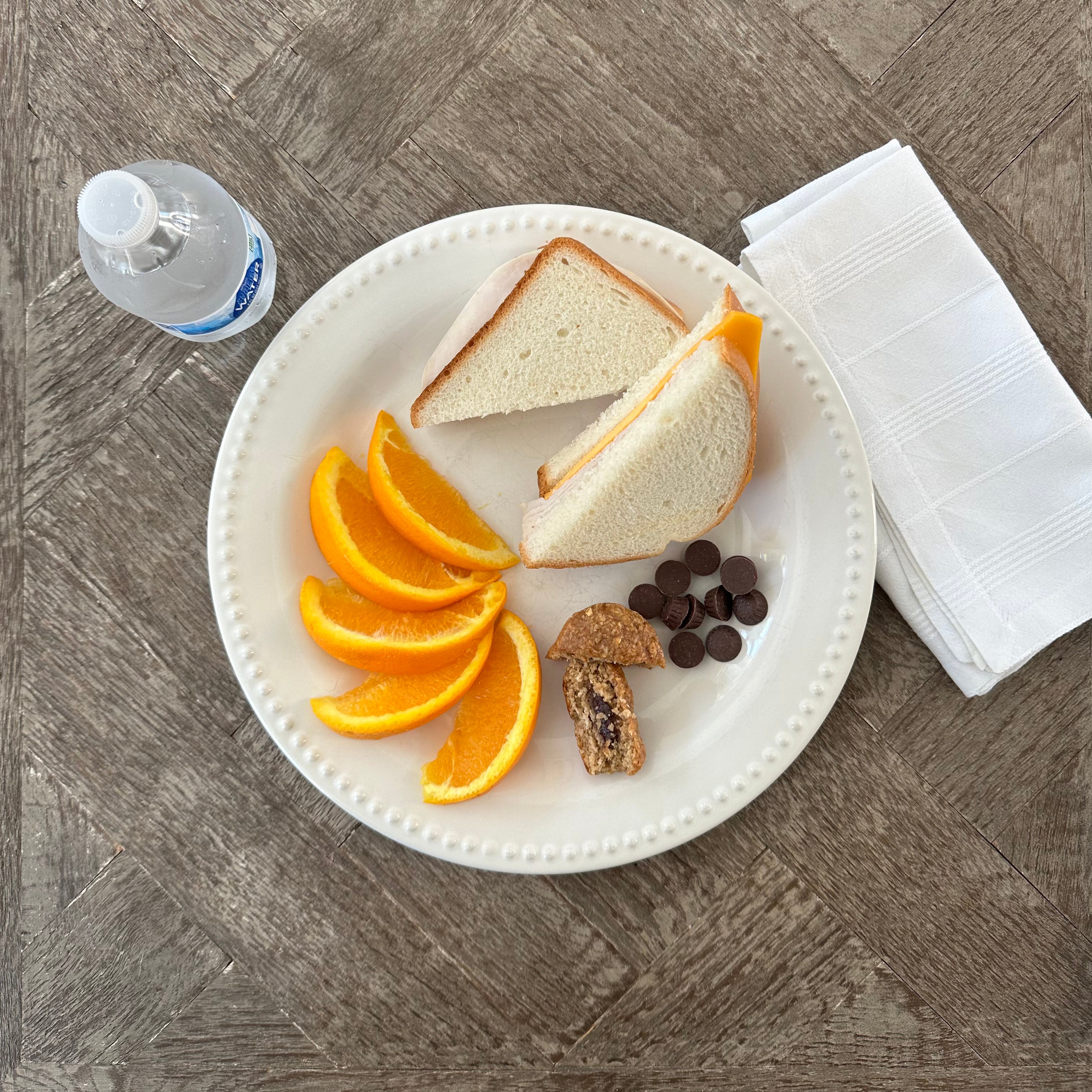 A plate with a Taylor Farms Turkey and Cheddar Sandwich on white bread, orange slices, a small piece of pastry, and chocolate pieces. A bottle of water and folded napkins are beside the plate on a wooden table.