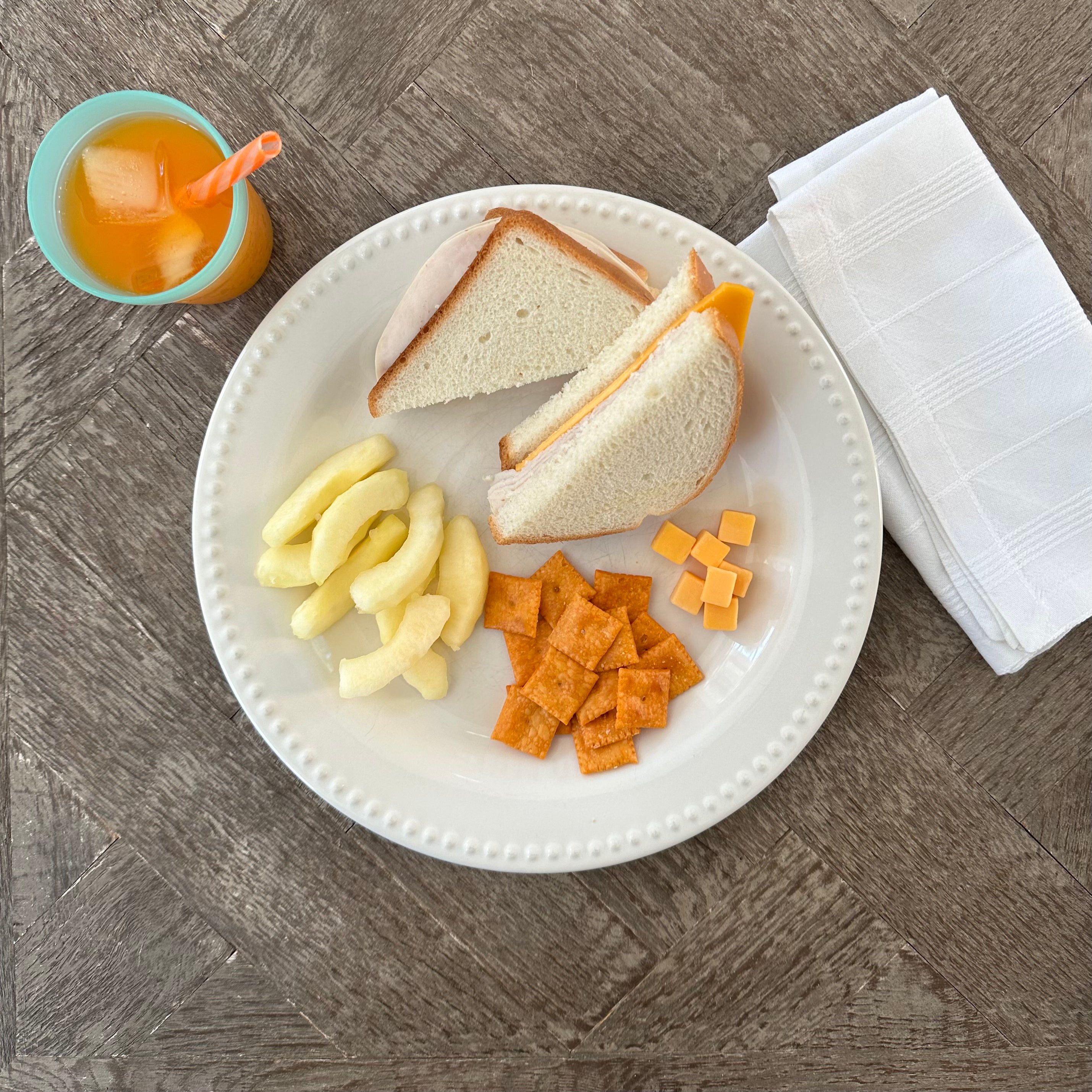 A plate with half a Turkey and Cheddar Sandwich by Taylor Farms, accompanied by apple slices, cheese crackers, and small mild cheddar cheese cubes rests on a wooden table. Beside it, there's a blue cup with an orange drink and a napkin.