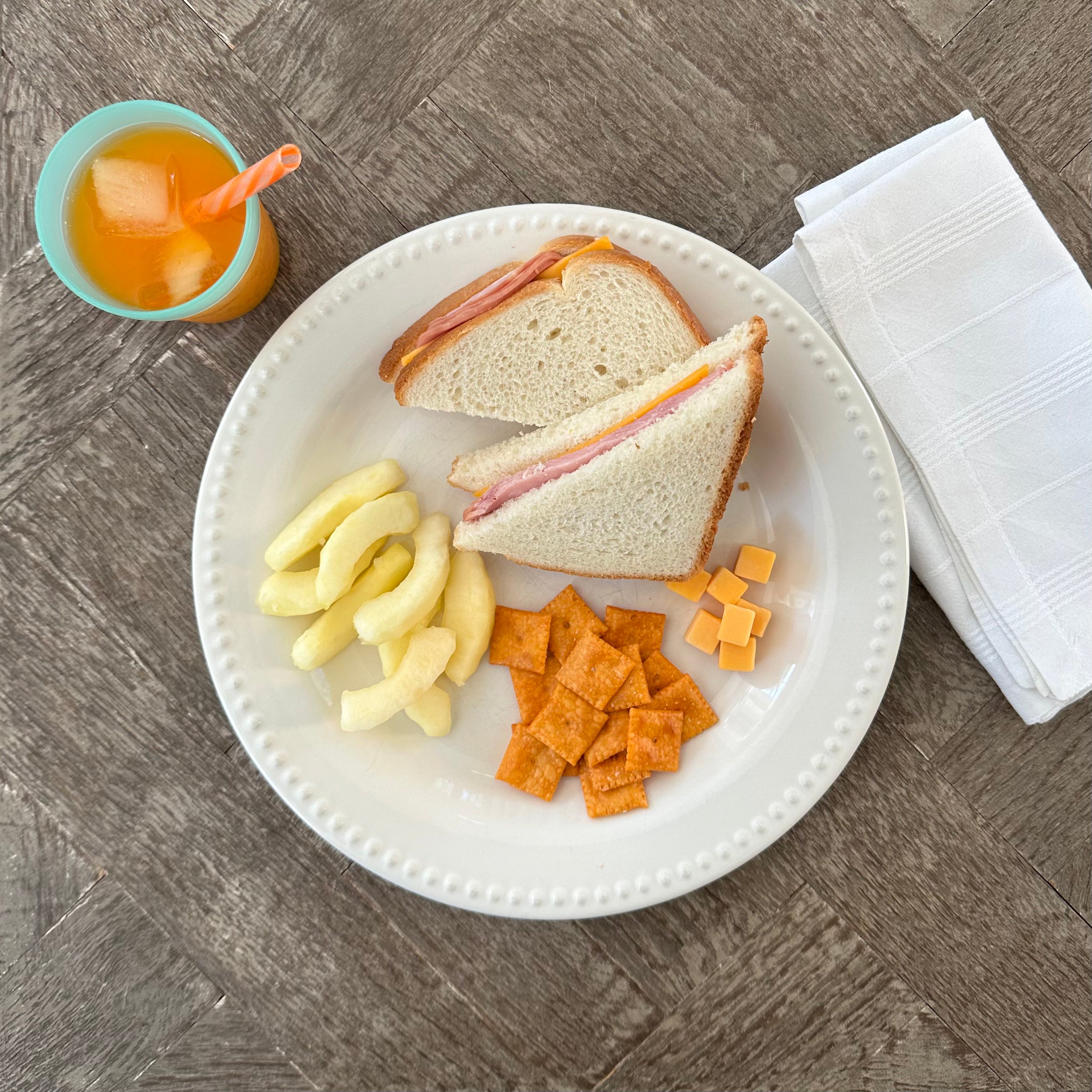 A plate with a Taylor Farms Ham and Cheddar Sandwich on white loaf bread, cheese crackers, puffed snacks, and cubed cheese. A blue cup with an orange drink and a striped straw is next to the plate, along with a folded white napkin.