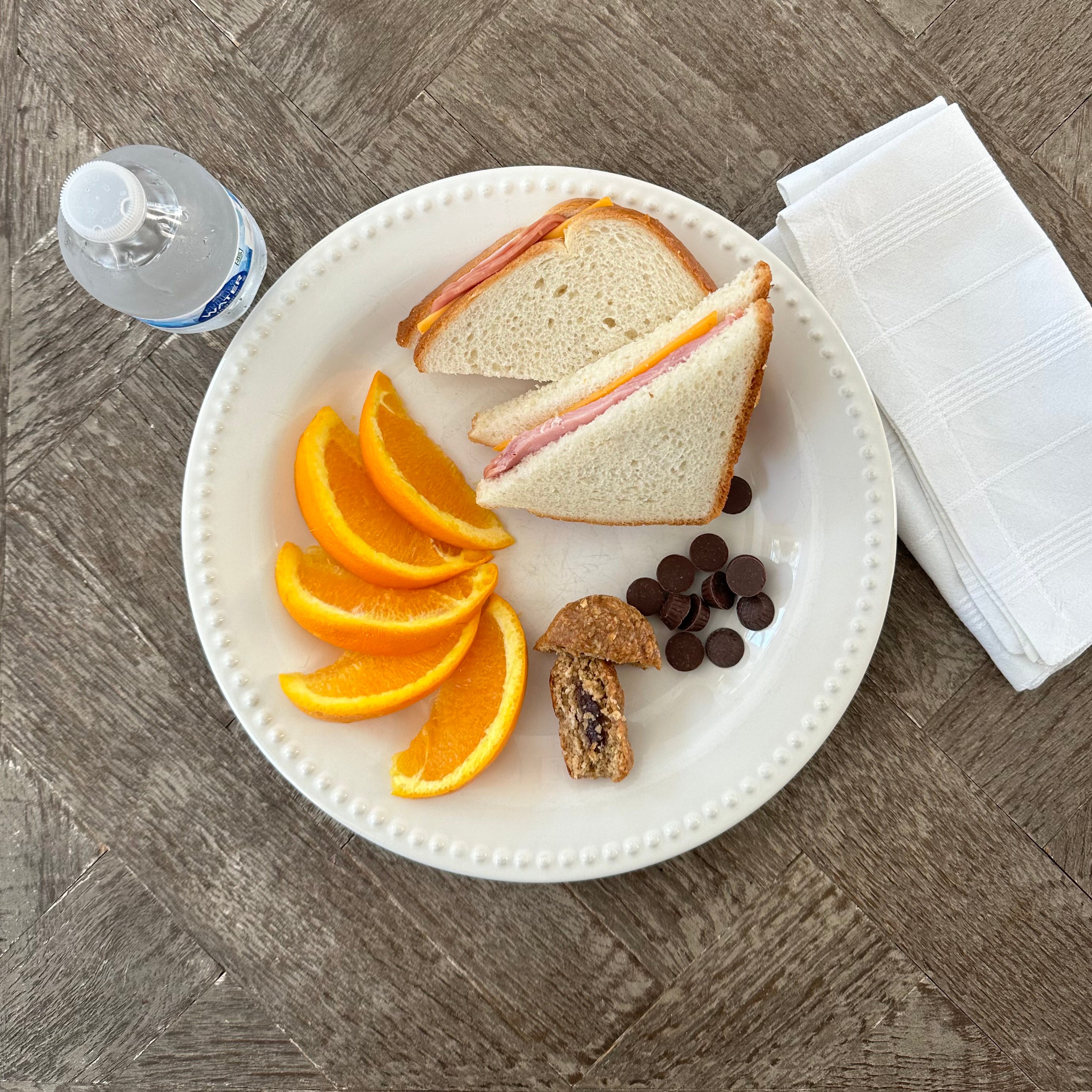 A plate featuring a Taylor Farms Ham and Cheddar Sandwich, alongside orange slices, chocolate chips, and a pastry. A bottle of water and a napkin are placed next to the plate on a wooden table.