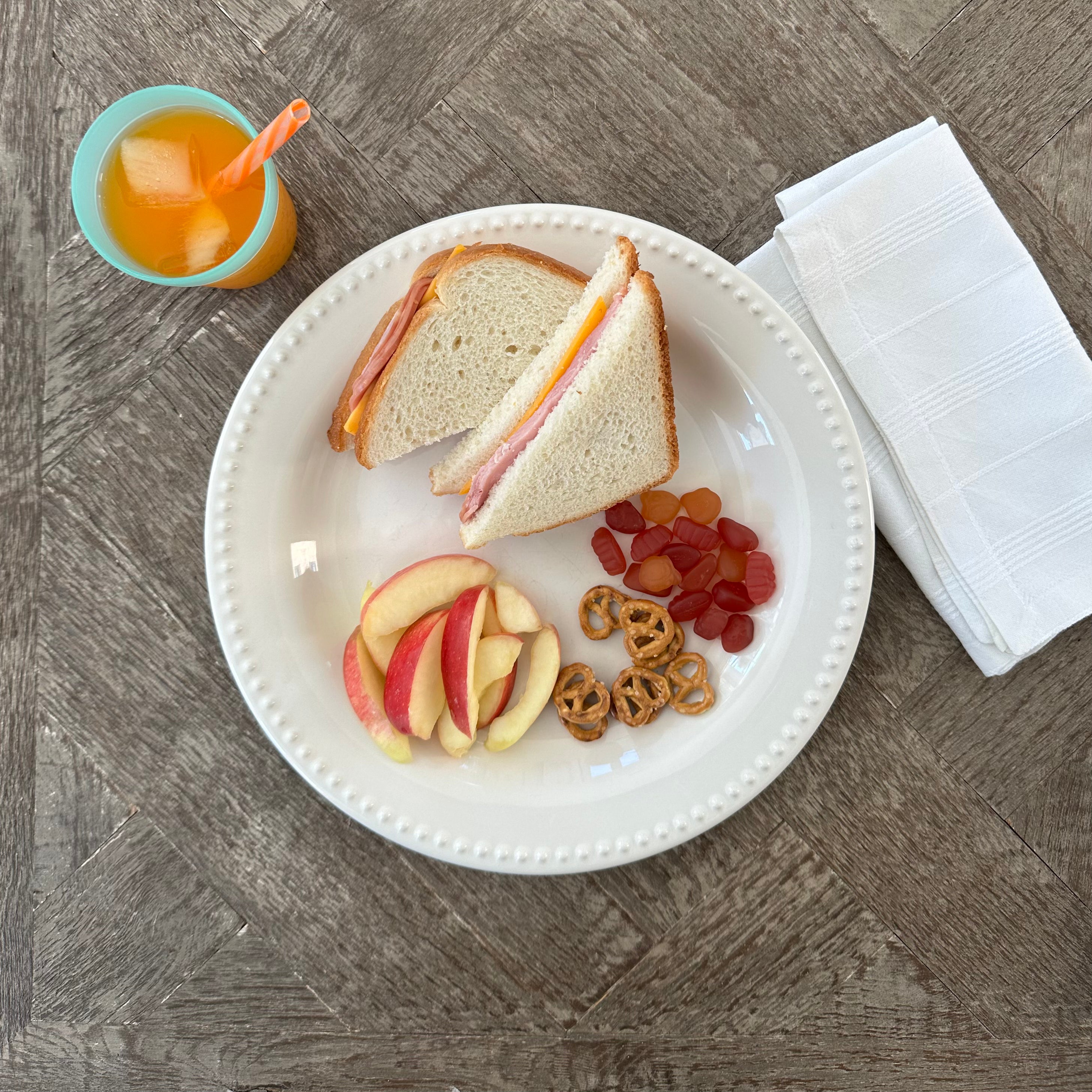 A wooden table features a plate with a Taylor Farms Ham and Cheddar Sandwich on white loaf bread, accompanied by apple slices, pretzels, and gummy candies. An orange juice cup with ice is placed next to a folded white napkin.