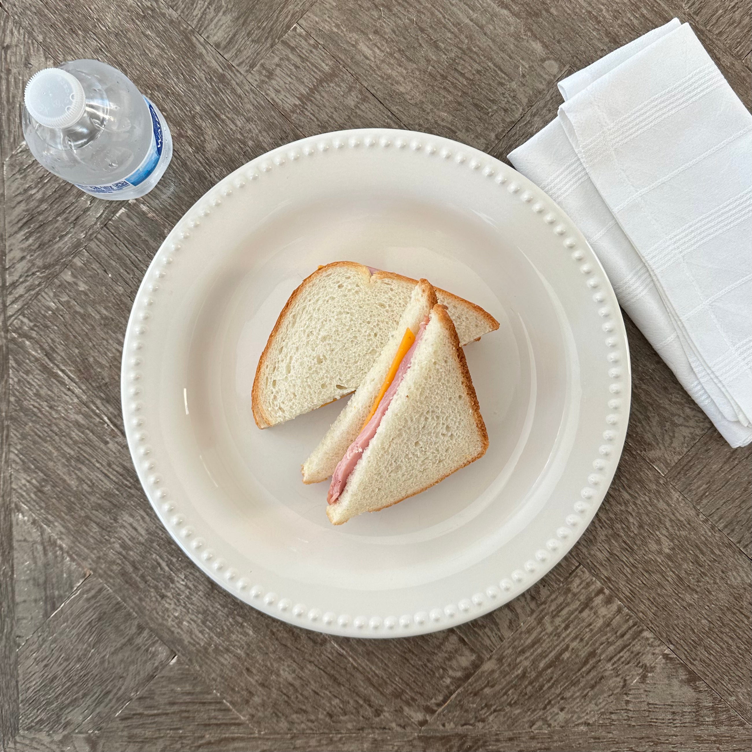 A Ham and Cheddar Sandwich from Taylor Farms sits on a white plate, placed next to a folded white napkin and a bottle of water on a wooden table.