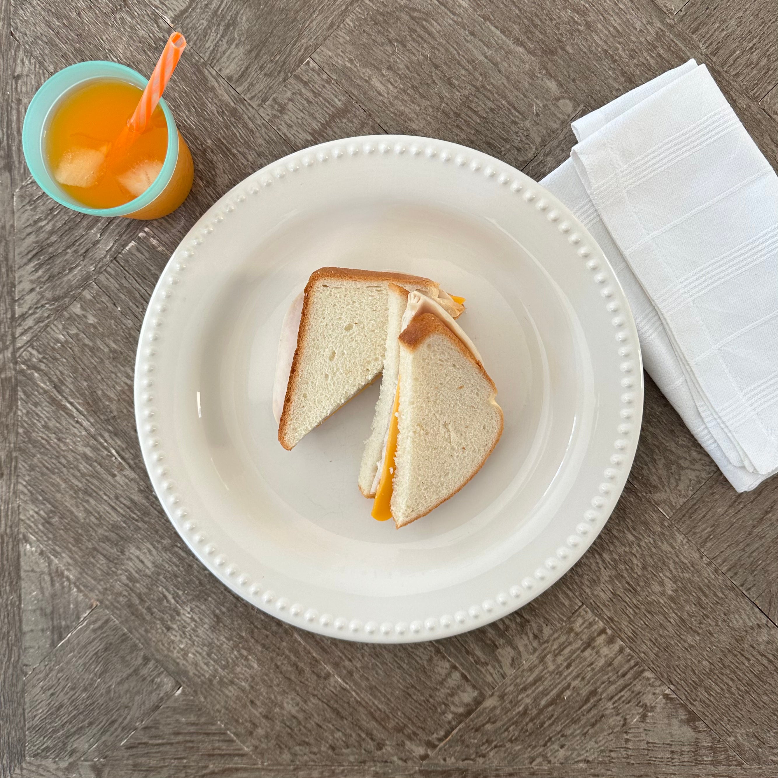 A Turkey and Cheddar Sandwich by Taylor Farms, cut in half on a white plate, sits beside a folded napkin and a cup of orange juice with a straw on a wooden table.