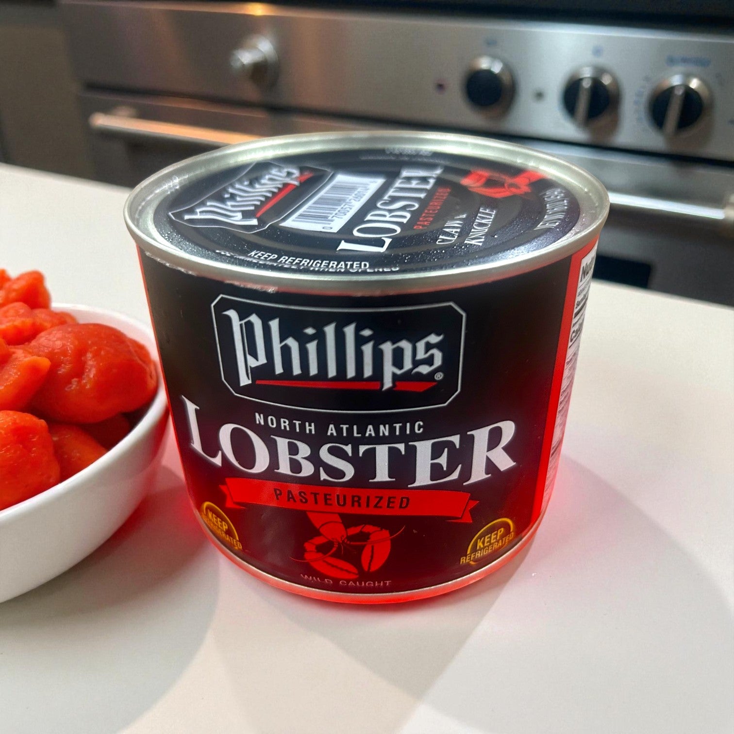 A can of Phillips Foods Lobster Meat – 16 Ounces sits on a countertop beside a bowl of orange food, with a stove in the background—premium ready-to-use seafood for your favorite recipes.