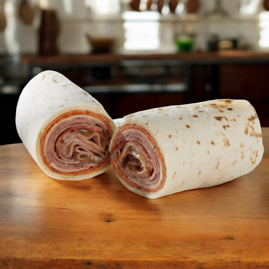Two E.A. Sween Italian Style Wraps, cut in half to display their deli meat and cheese layers, rest on a wooden surface against a blurred kitchen backdrop, embodying authentic Italian cuisine. Ships frozen; keeps 30 days in the cooler once thawed.